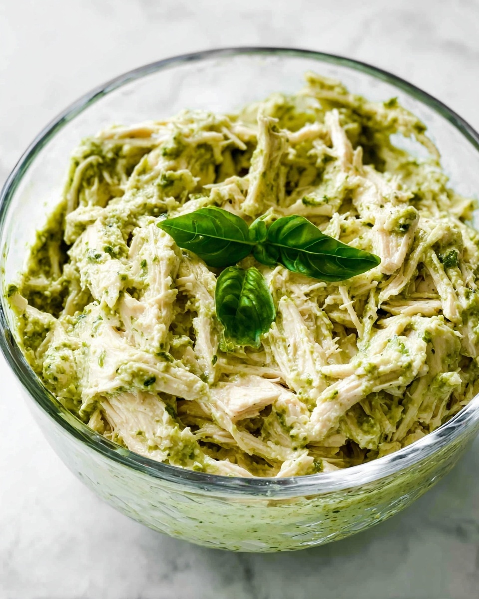 A clear glass bowl contains a mix of shredded light-colored chicken and creamy green pesto sauce. The mixture has a thick and slightly chunky texture, with visible strands of chicken coated in a smooth green pesto. On top, two fresh bright green basil leaves add a touch of color contrast. The bowl is placed on a surface with a white marbled texture. photo taken with an iphone --ar 4:5 --v 7