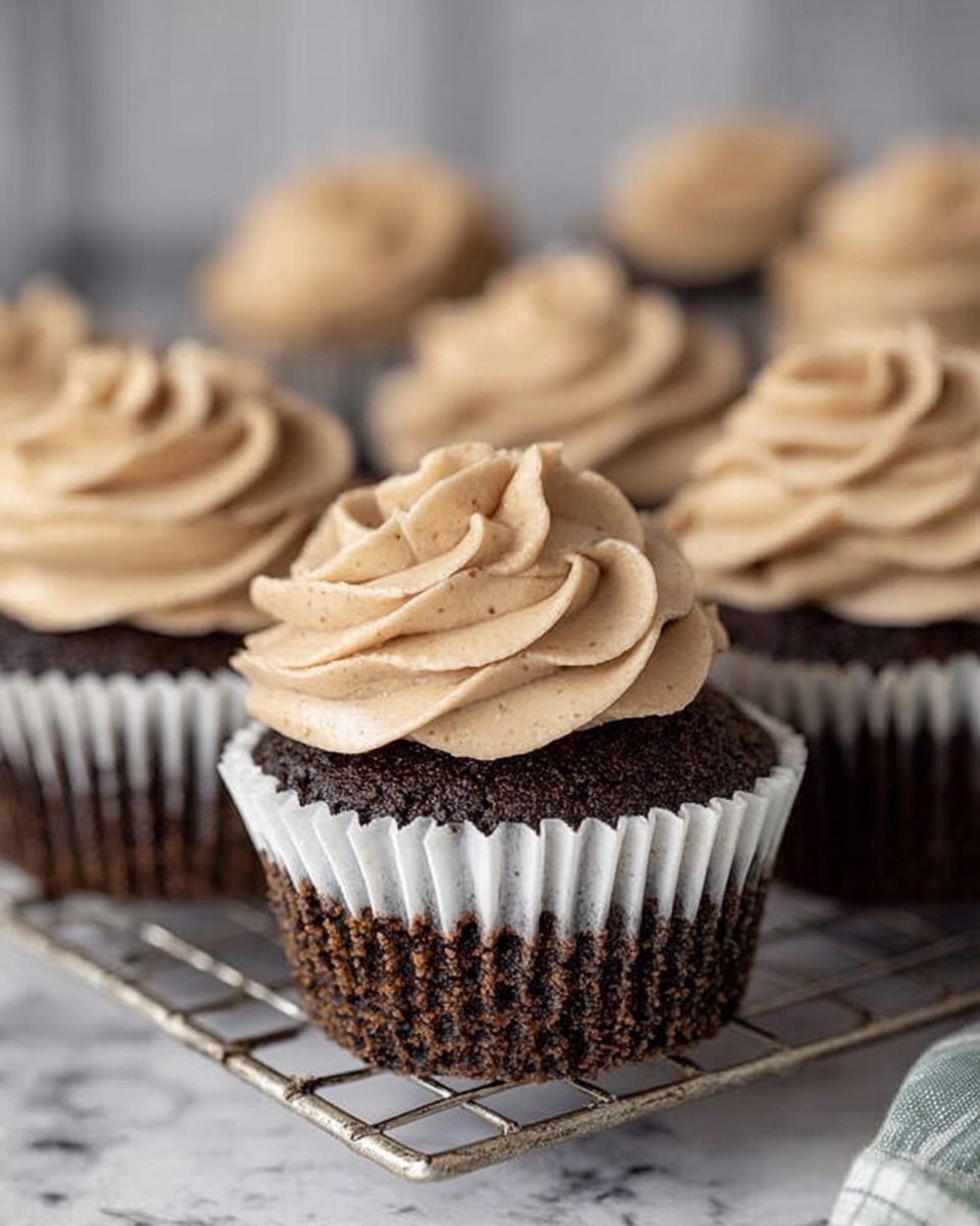 A group of chocolate cupcakes arranged on a silver cooling rack over a white marbled surface, each cupcake wrapped in a white paper liner with dark chocolate crumbs visible on the liner. Each cupcake has one thick layer of creamy light brown frosting piped in a floral swirl on top with a soft, smooth texture and tiny specks running through it. The chocolate cake base looks moist and dark, creating a rich contrast with the frosting above. The view focuses closely on one cupcake in front with others softly blurred in the background. Photo taken with an iphone --ar 4:5 --v 7