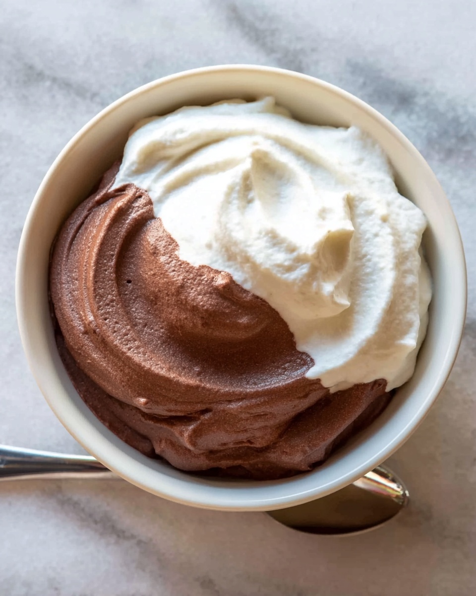 A white bowl with two layers of smooth mousse placed on a white marbled surface. The bottom layer is rich dark brown chocolate mousse with a creamy texture and slight swirls on top. The second layer is a thick soft white vanilla mousse sitting on the chocolate mousse, showing a smooth uneven surface and gentle folds. A shiny silver spoon is placed next to the bowl, partially visible. photo taken with an iphone --ar 4:5 --v 7