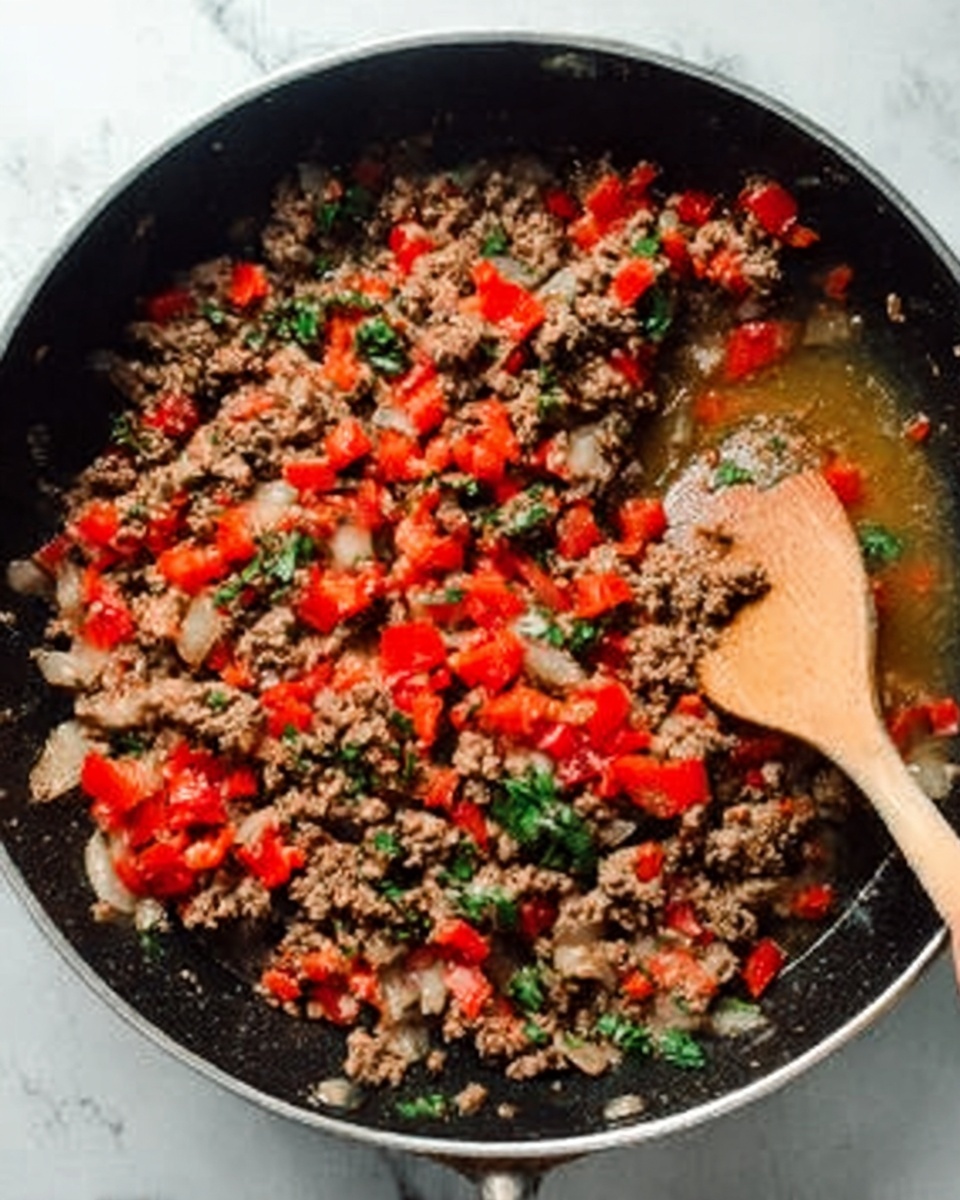 The image shows a black pan filled with a cooked mix of ground meat, chopped red bell peppers, onions, and green herbs. The ground meat is brown and crumbly, spread evenly across the pan. Bright red pieces of bell pepper are scattered throughout, adding pops of color that contrast with the light, translucent onion slices. Green leafy herbs are mixed in, giving a fresh look to the dish. A woman's hand holds a wooden spoon resting inside the pan, positioned near the top right. The background is a white marbled surface. Photo taken with an iphone --ar 4:5 --v 7