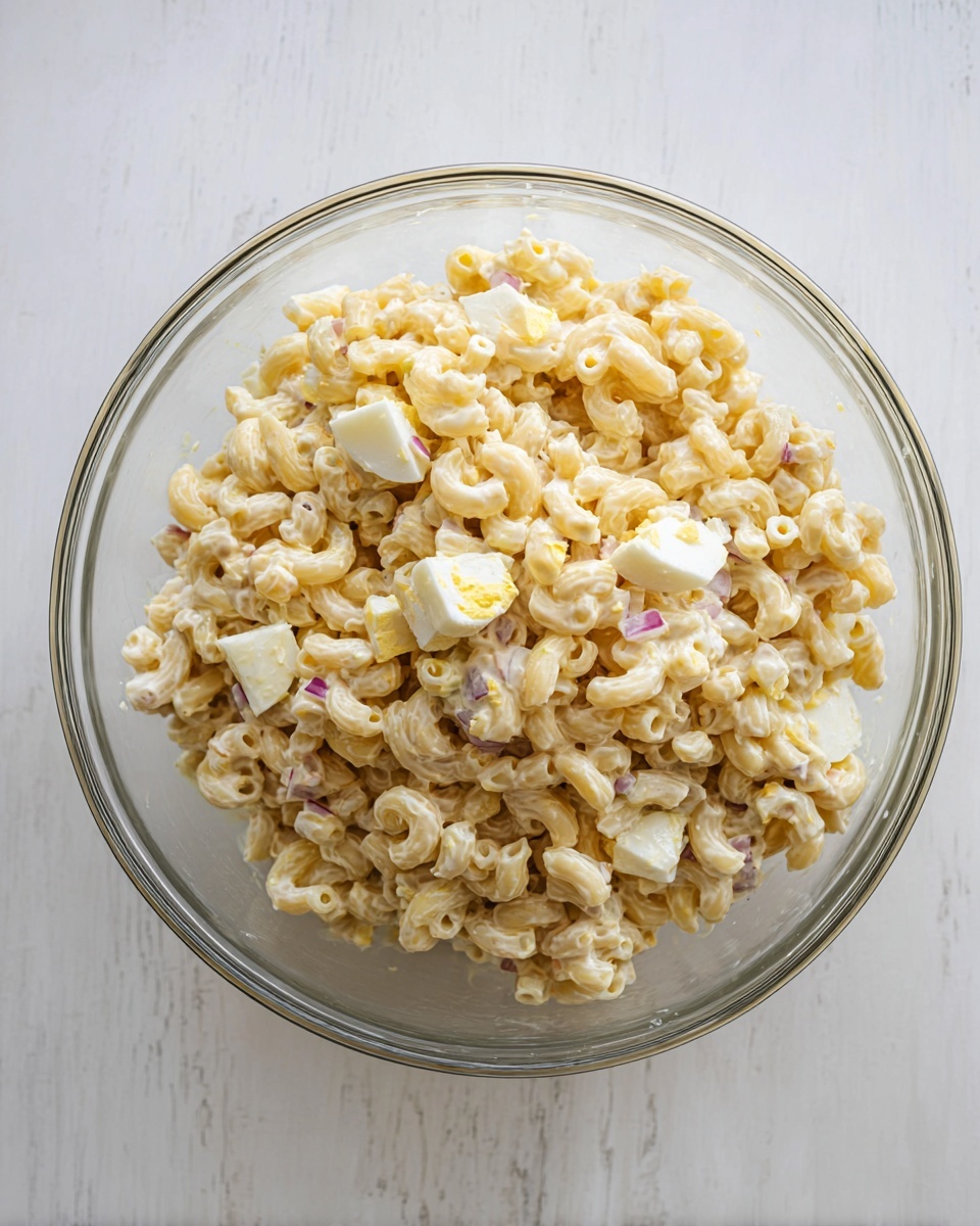 A clear glass bowl filled with a creamy macaroni salad sits on a white marbled surface. The salad has one main layer made of small curved macaroni pieces coated in a thick, pale yellow dressing, mixed evenly with small chunks of hard-boiled egg white and yolk, and tiny bits of purple onion scattered throughout. The textures vary slightly with the smooth pasta and soft egg pieces. The bowl is shown from above, highlighting the mix inside. Photo taken with an iphone --ar 4:5 --v 7