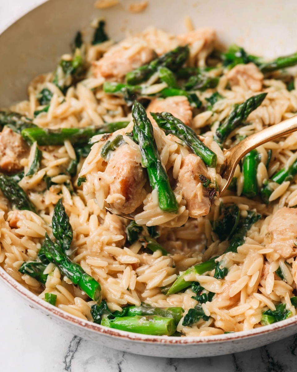 A close-up view of a white pan filled with a creamy dish made of small, light beige orzo pasta mixed with light golden pieces of chicken and vibrant green asparagus pieces. The asparagus is cut into medium-sized segments with a textured, slightly crisp look. The sauce is smooth and creamy, coating all the ingredients evenly, giving the dish a rich and glossy texture. A gold spoon is scooping a portion of the dish, showing the mix of orzo, chicken, and asparagus clearly. The background has a white marbled texture. Photo taken with an iphone --ar 4:5 --v 7