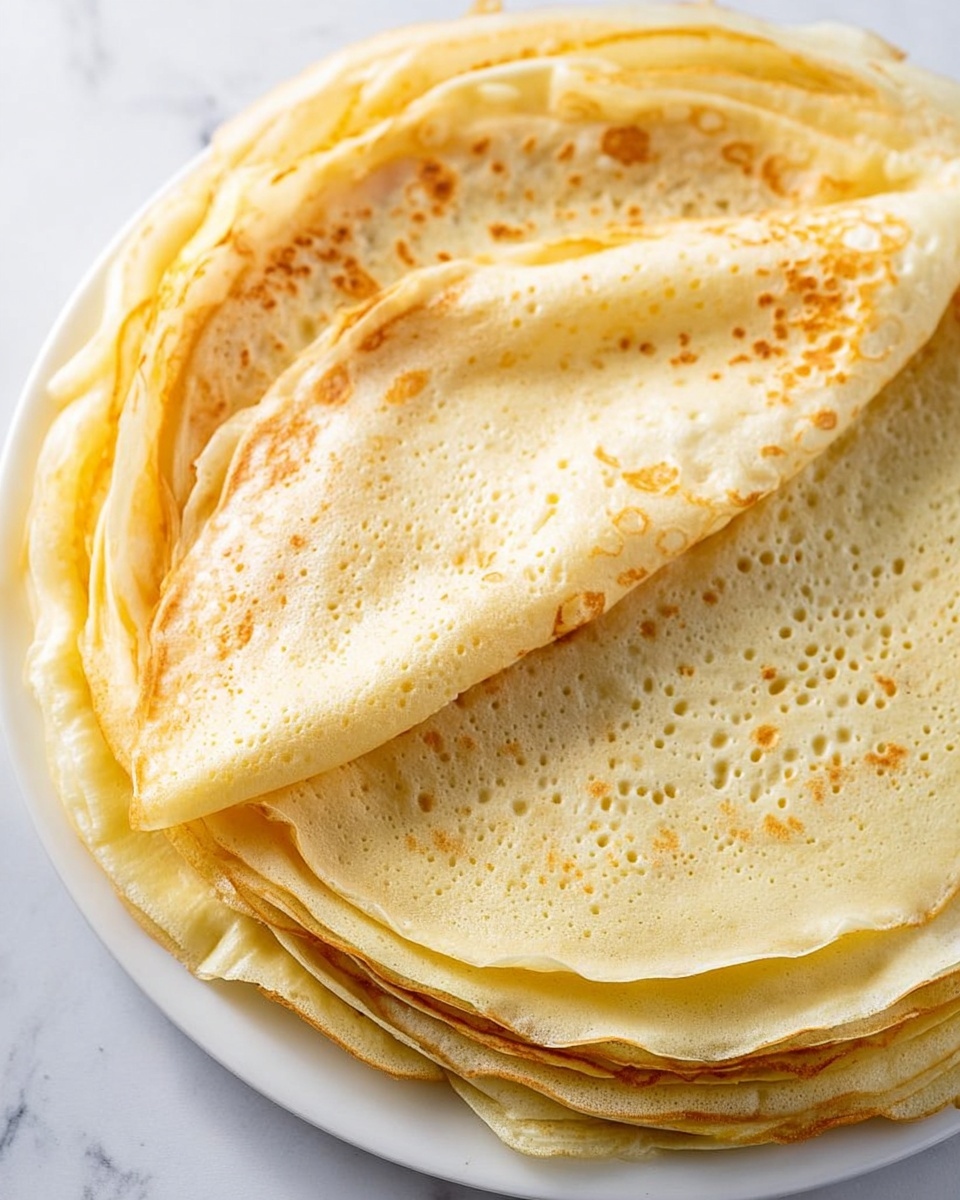A stack of thin crepes is shown on a white plate placed on a white marbled surface. There are many crepes layered on top of each other, each crepe pale yellow with light golden brown spots and edges. The top crepe is partially folded over, showing its soft and smooth texture with small air bubbles and tiny holes across the surface. The crepes look light and delicate with slightly uneven edges. photo taken with an iphone --ar 4:5 --v 7