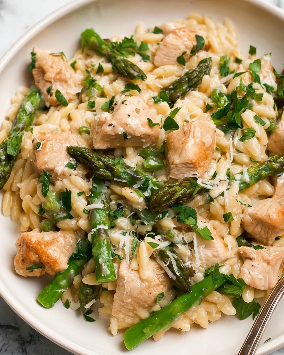 A close-up view of a creamy pasta dish on a white plate, featuring three main layers: chunks of light golden chicken pieces scattered throughout, bright green asparagus cut into large pieces, and a creamy sauce coating small, rice-shaped pasta. The dish is garnished with finely chopped fresh parsley and grated white cheese sprinkled on top, adding texture and color contrast. The background shows a white marbled texture surface, and a silver fork is partially visible on the right side of the plate. photo taken with an iphone --ar 4:5 --v 7