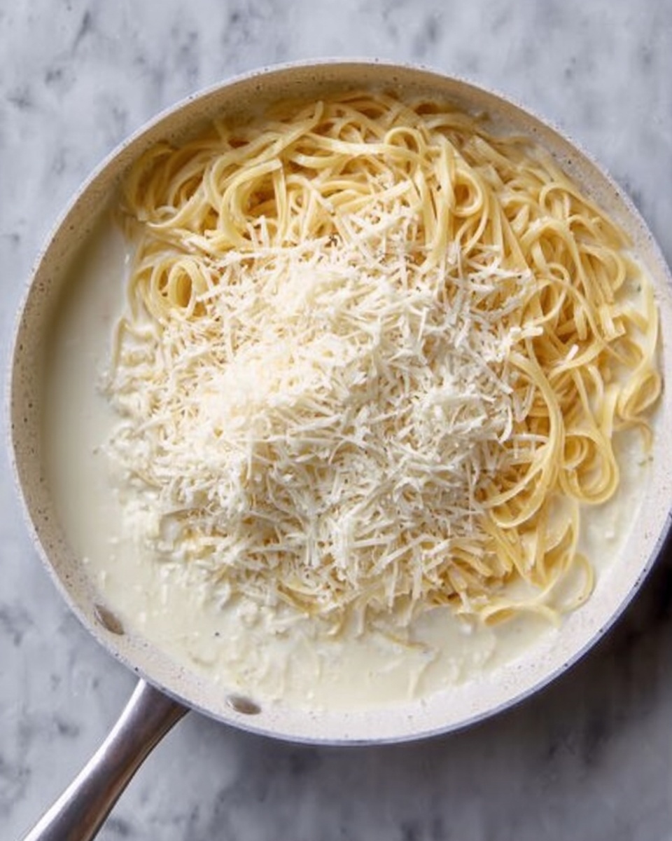 A white pan holds creamy white sauce covering half of light yellow pasta noodles, with a generous pile of finely grated white cheese over the center. The pan is on a white marbled surface. Photo taken with an iphone --ar 4:5 --v 7