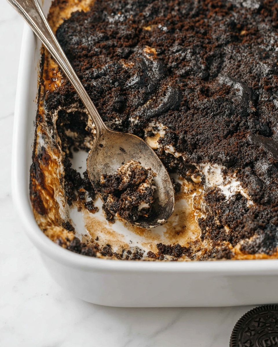 The image shows a white baking dish with a thick layer of dark brown and black crumbly topping, uneven in texture with some rough edges and burnt spots around the sides. Under the top layer, there are hints of light tan and creamy areas, suggesting melted marshmallows or a similar gooey layer. A vintage metal spoon with a worn surface rests inside the dish, with part of the topping scooped out, revealing the white dish underneath. The dish sits on a white marbled surface, and a partial Oreo cookie is visible at the bottom right corner. photo taken with an iphone --ar 4:5 --v 7
