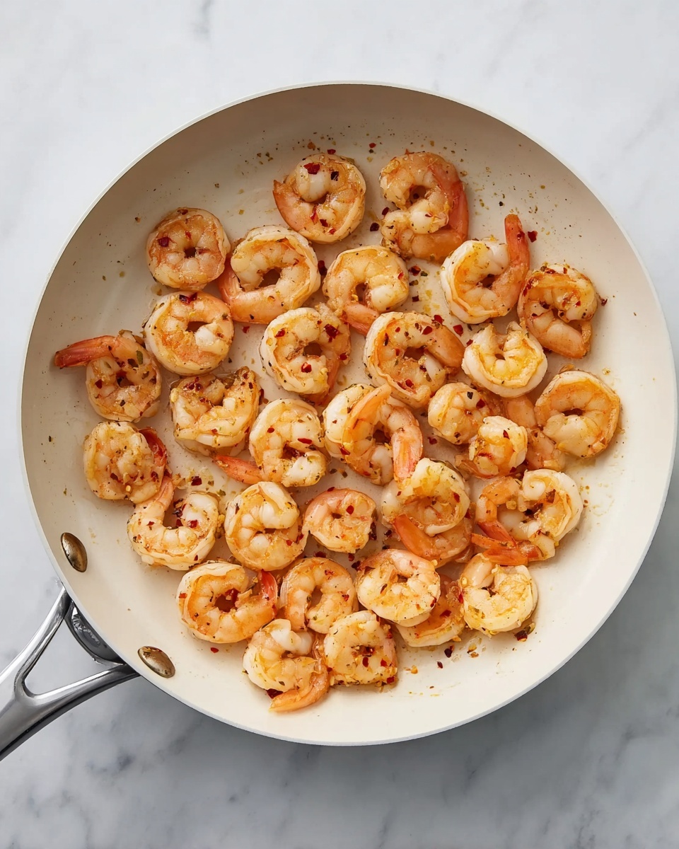 The image shows a gray frying pan filled with cooked shrimp. The shrimp are light orange with some reddish spots from seasoning and are spread evenly around the pan. The pan sits on a white marbled surface, and the shrimp have a slightly shiny, cooked texture. The metal handle of the pan is visible on the left side. photo taken with an iphone --ar 4:5 --v 7