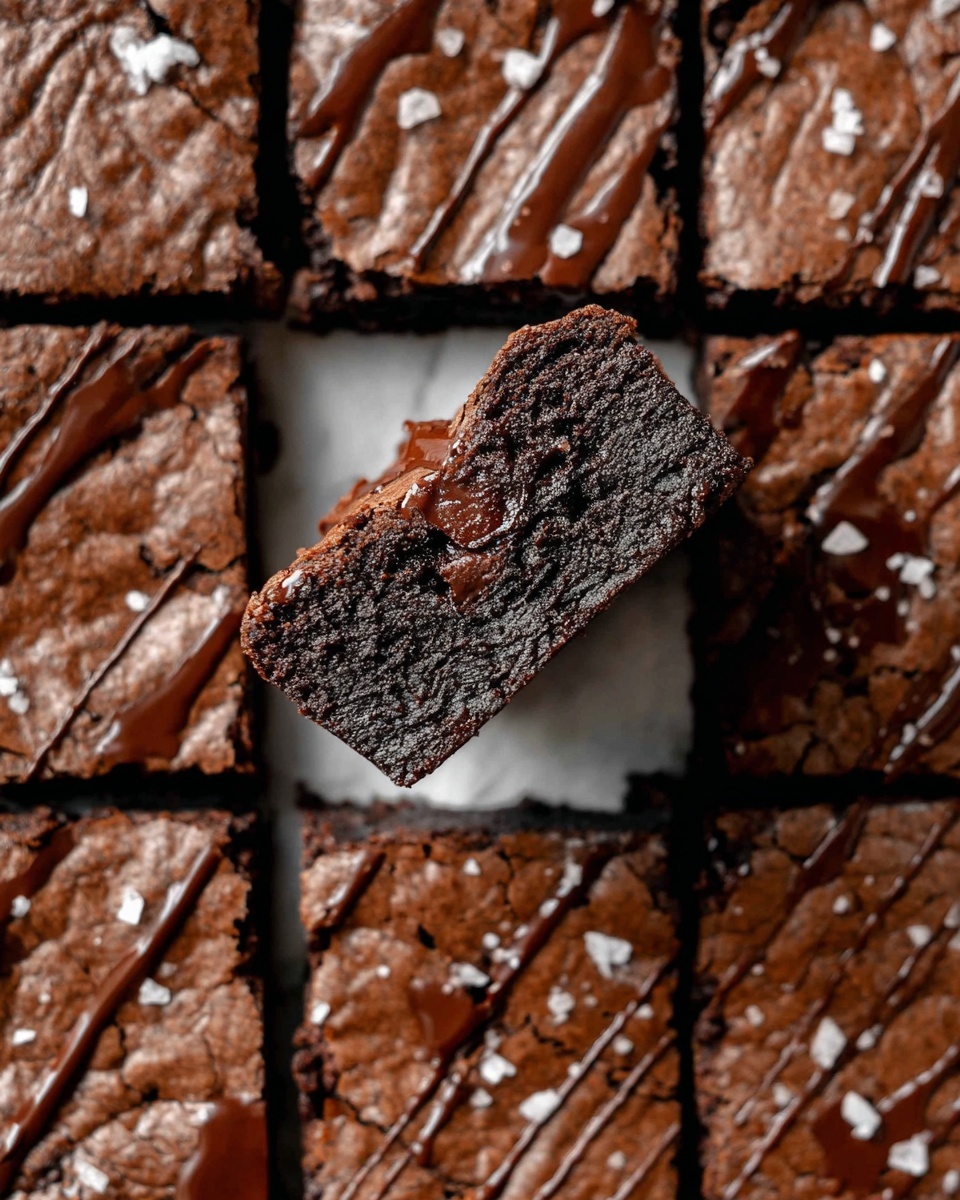 A tray of dark brown chocolate brownies is displayed on a white marbled surface, each square topped with a drizzle of darker chocolate and scattered sea salt flakes. One brownie square is held above the tray by a woman's hand, showing the inside texture with visible melted chocolate chunks, dense and moist with a slightly shiny surface. The edges of the brownies are cleanly cut, and the tray is arranged in a grid with one square missing where the held piece was taken. Photo taken with an iphone --ar 4:5 --v 7