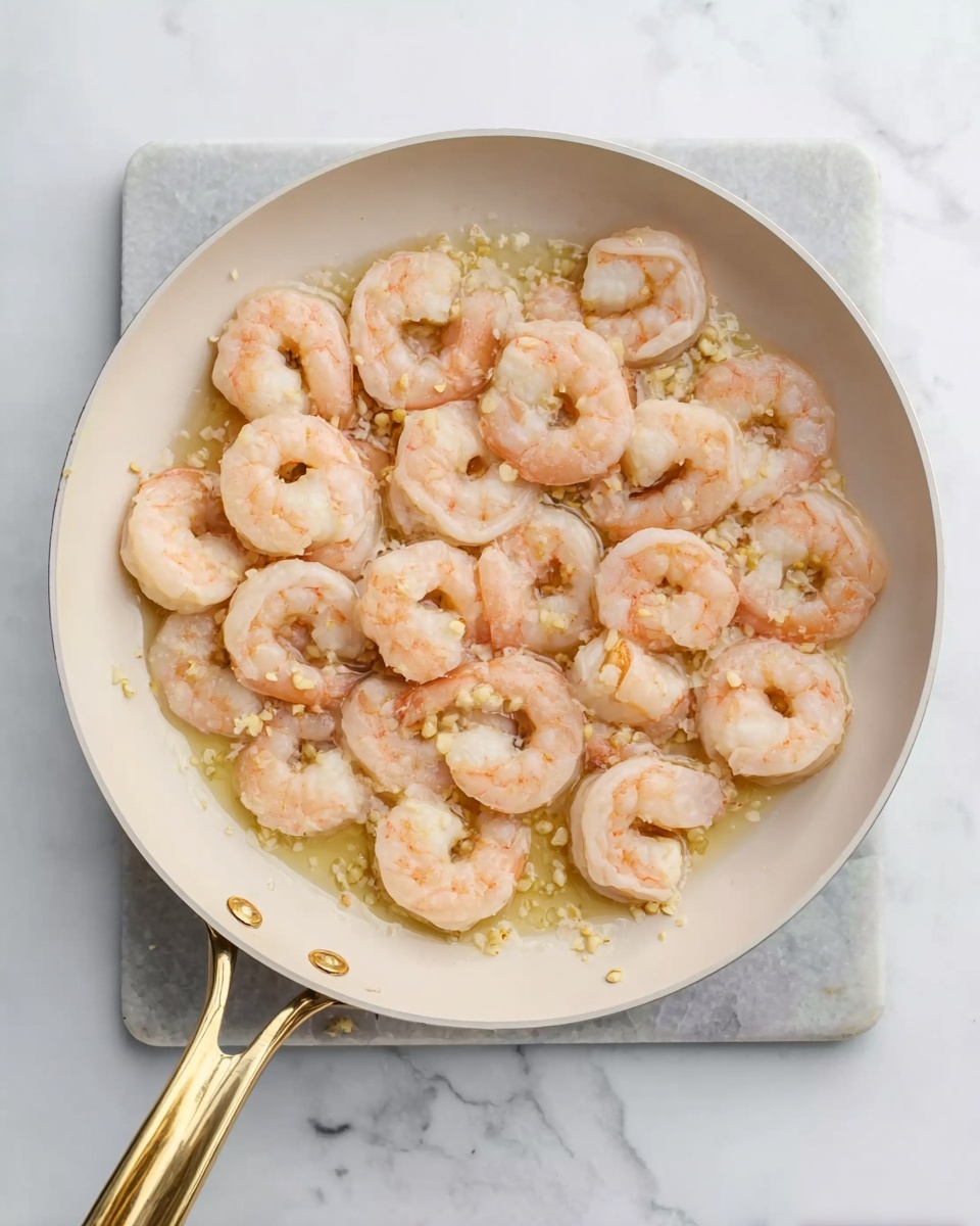 A top view of a white pan with a gold handle filled with evenly placed cooked shrimp. The shrimp are pink and white with a slight glossy texture, sitting in a light garlic butter sauce that has small bits of garlic visible throughout. The pan rests on a white marbled surface that adds a clean and bright background to the image. photo taken with an iphone --ar 4:5 --v 7
