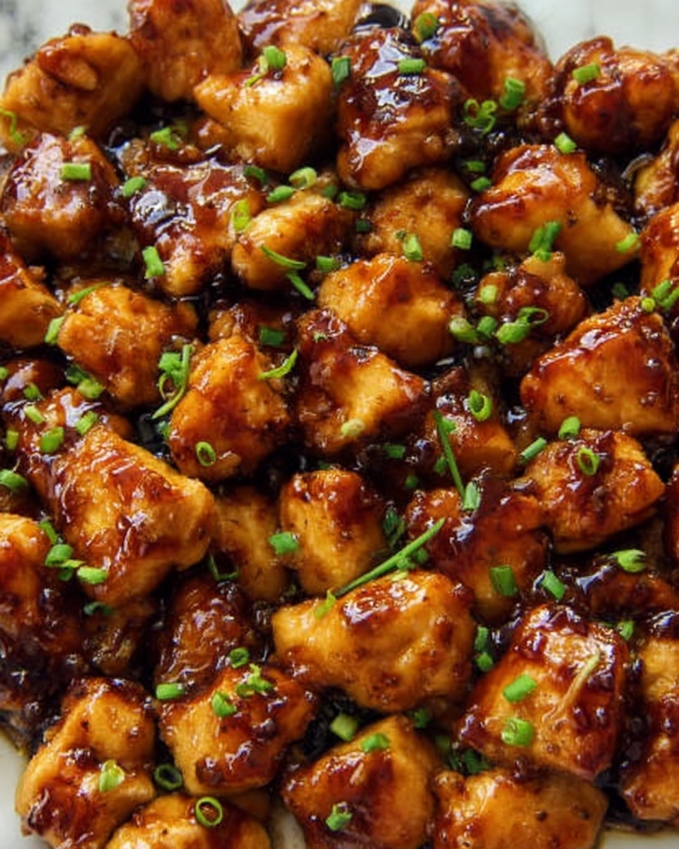 The image shows many pieces of cooked chicken with a shiny dark brown sauce on them, making them look glazed and juicy. Small green chopped bits are sprinkled on top, adding color contrast. The texture of the chicken pieces looks soft and slightly crisp on the outside. The dish is shown close-up on a white marbled surface. photo taken with an iphone --ar 4:5 --v 7