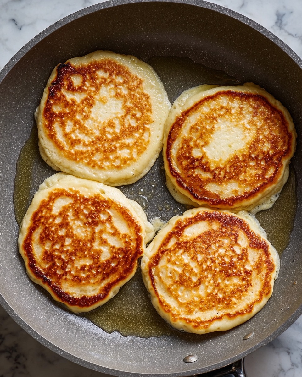The image shows four thick pancakes cooking in a grey frying pan. Each pancake is golden brown with a light, bubbly texture on the surface, and slightly crispy edges that are darker in color. The pancakes are round but not perfectly shaped, with some unevenness around the sides where the batter spread. The pan has a shiny layer of oil around the pancakes, catching the light and adding a glossy look. The background has a white marbled texture. photo taken with an iphone --ar 4:5 --v 7