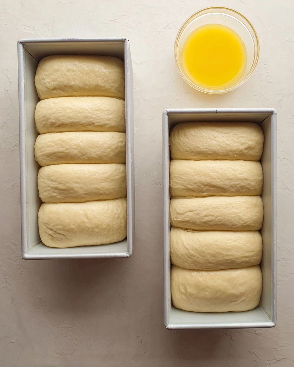 Two white metal loaf pans are placed side by side on a white marbled surface. Each pan holds soft, pale dough arranged in six rolled pieces, stacked neatly to fill the pans. The dough has a smooth texture with a slightly uneven surface, pale yellow color, and looks ready to rise or bake. To the right of the pans, there is a small clear container filled with a bright yellow liquid, likely egg wash or butter. The scene is simple and clean, showing an early stage of bread preparation. photo taken with an iphone --ar 4:5 --v 7
