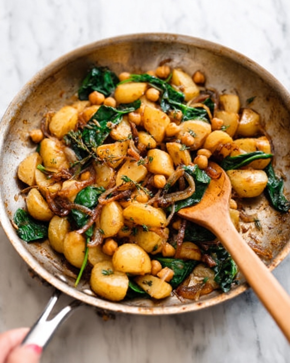 In a stainless steel pan on a white marbled surface, there are small pieces of golden browned potatoes mixed with bright green spinach leaves. Light brown small round chickpeas are scattered throughout, along with pieces of caramelized onions and some herbs on top. A wooden spoon is stirring the mix, held by a woman's hand at the edge of the pan. The colors are warm and inviting, with a mix of crispy and soft textures visible in the cooked ingredients. Photo taken with an iphone --ar 4:5 --v 7