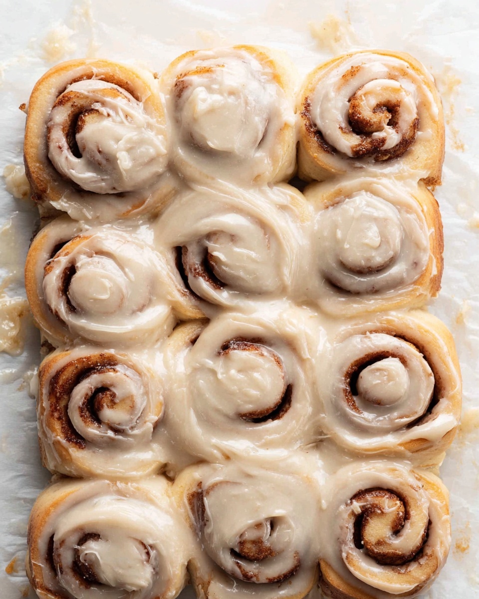The image shows a group of nine cinnamon rolls placed tightly together in a 3 by 3 arrangement on white parchment paper. Each cinnamon roll has a visible spiral pattern with a light golden brown dough color and darker brown cinnamon filling. The rolls are generously covered with a thick layer of creamy, beige-colored frosting that is smooth and slightly shiny, dripping over the edges and pooling a bit on the parchment paper. The texture of the dough looks soft and fluffy beneath the frosting. The background is a white marbled texture. photo taken with an iphone --ar 4:5 --v 7