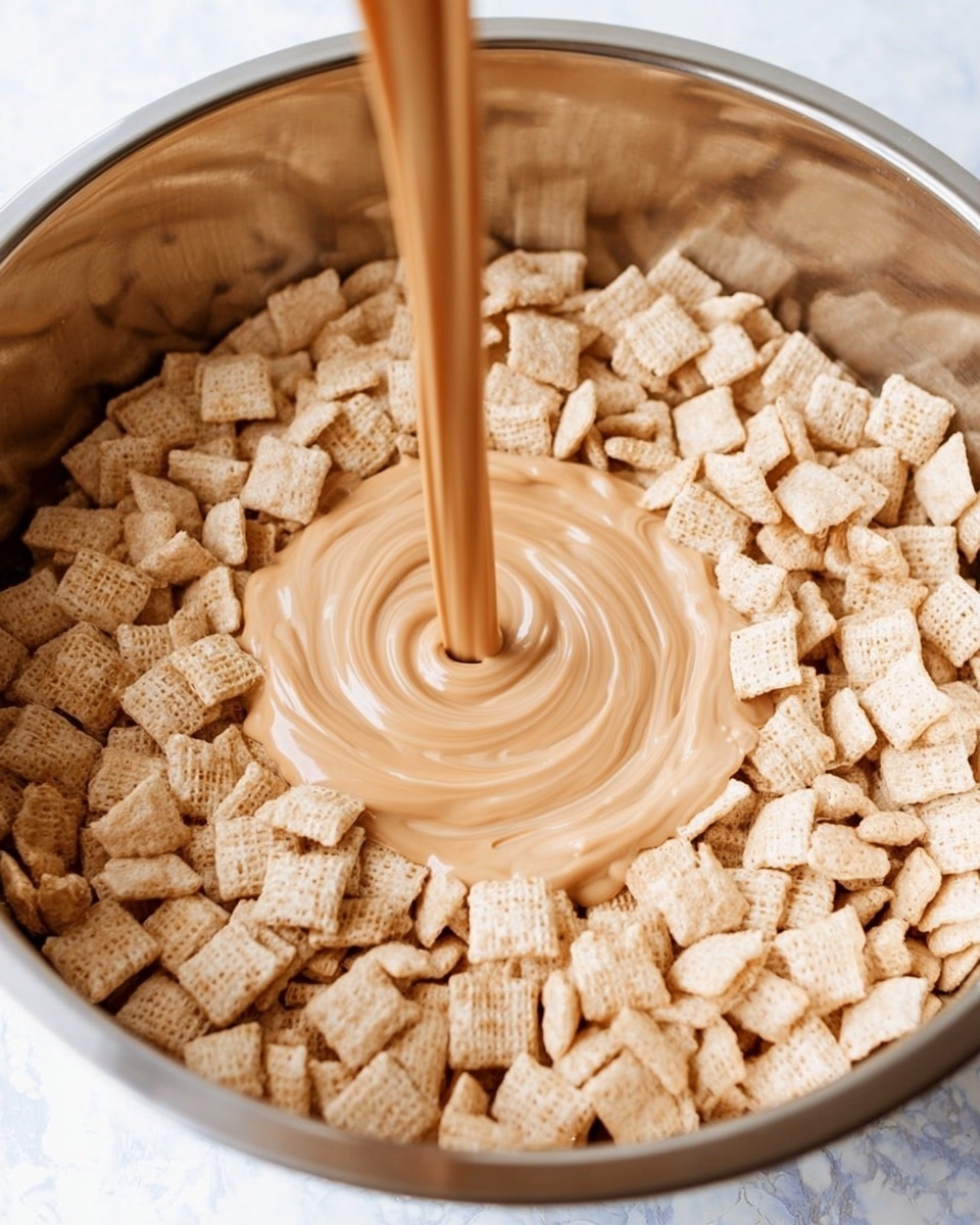 A large metal bowl is filled with a single layer of small square cereal pieces that are light beige in color with a slightly rough texture. A stream of smooth, light brown creamy mixture is being poured into the center of the cereal, creating a spiral shape on top of the squares. The background surface is white with a marbled texture. photo taken with an iphone --ar 4:5 --v 7