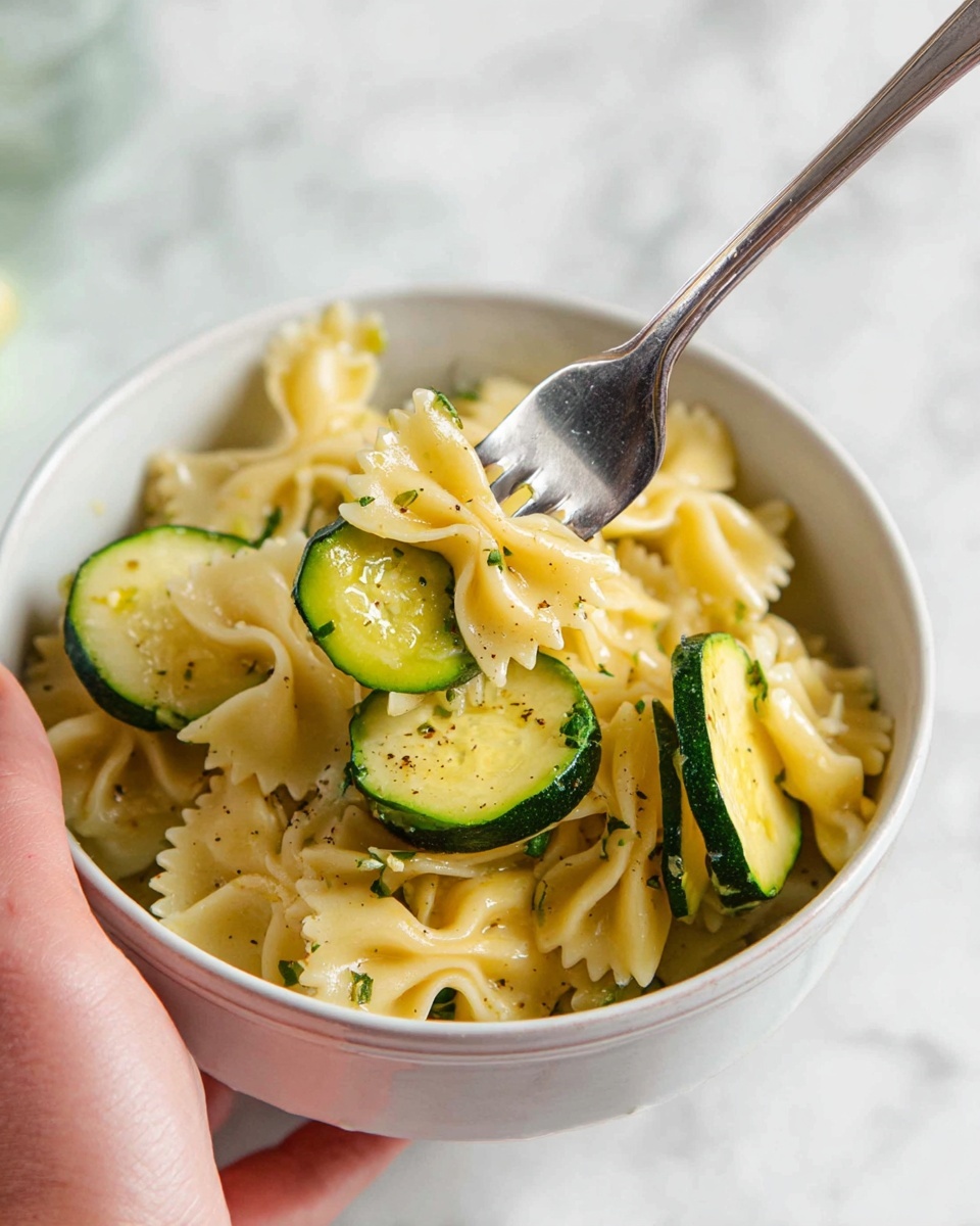 A white bowl filled with light yellow bowtie pasta mixed with thick green zucchini slices. The zucchini pieces are on top and around the pasta, showing a slightly shiny texture. A woman's hand holds a silver fork near the center, spearing a piece of zucchini and some pasta. The background is a soft white marbled surface, giving a clean and simple look. photo taken with an iphone --ar 4:5 --v 7