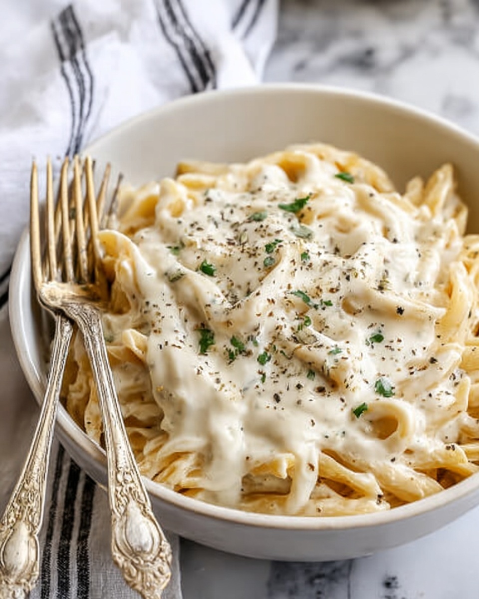 A white bowl filled with pasta topped generously with creamy white sauce that covers most of the pasta, sprinkled with small bits of green herbs and black pepper on top, showcasing the textures of the sauce and pasta clearly. Two ornate forks rest on the side of the bowl on a white marbled surface with a white cloth with black stripes partly visible in the background. Photo taken with an iphone --ar 4:5 --v 7