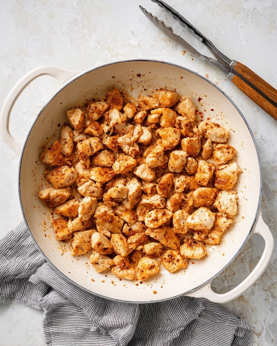 A white pan filled with many small, browned chicken pieces that have a light golden color with some darker brown spots and a slightly rough texture. The chicken pieces are spread evenly across the pan’s flat surface. The pan has two handles and is resting on a white marbled surface with a gray and white striped cloth partially underneath it on the left side. A pair of metal tongs with wooden grips lies on the top right corner of the image. photo taken with an iphone --ar 4:5 --v 7