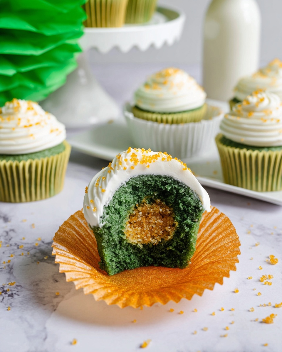 The image shows green cupcakes with three main layers. The bottom layer is a round green cupcake with a spongy texture inside a shiny orange cupcake liner. The middle layer inside the cupcake shows a hollow filled with golden sugar crystals that sparkle. The top layer is a smooth swirl of white frosting dotted with more golden sugar sprinkles. One cupcake is cut in half and placed in a white cupcake liner, revealing the golden crystal center. In the background, more whole cupcakes sit on a white marbled surface with a white two-tiered tray and a white bottle partly visible. The scene is bright and clean, with some green paper decorations adding extra color. Photo taken with an iphone --ar 4:5 --v 7