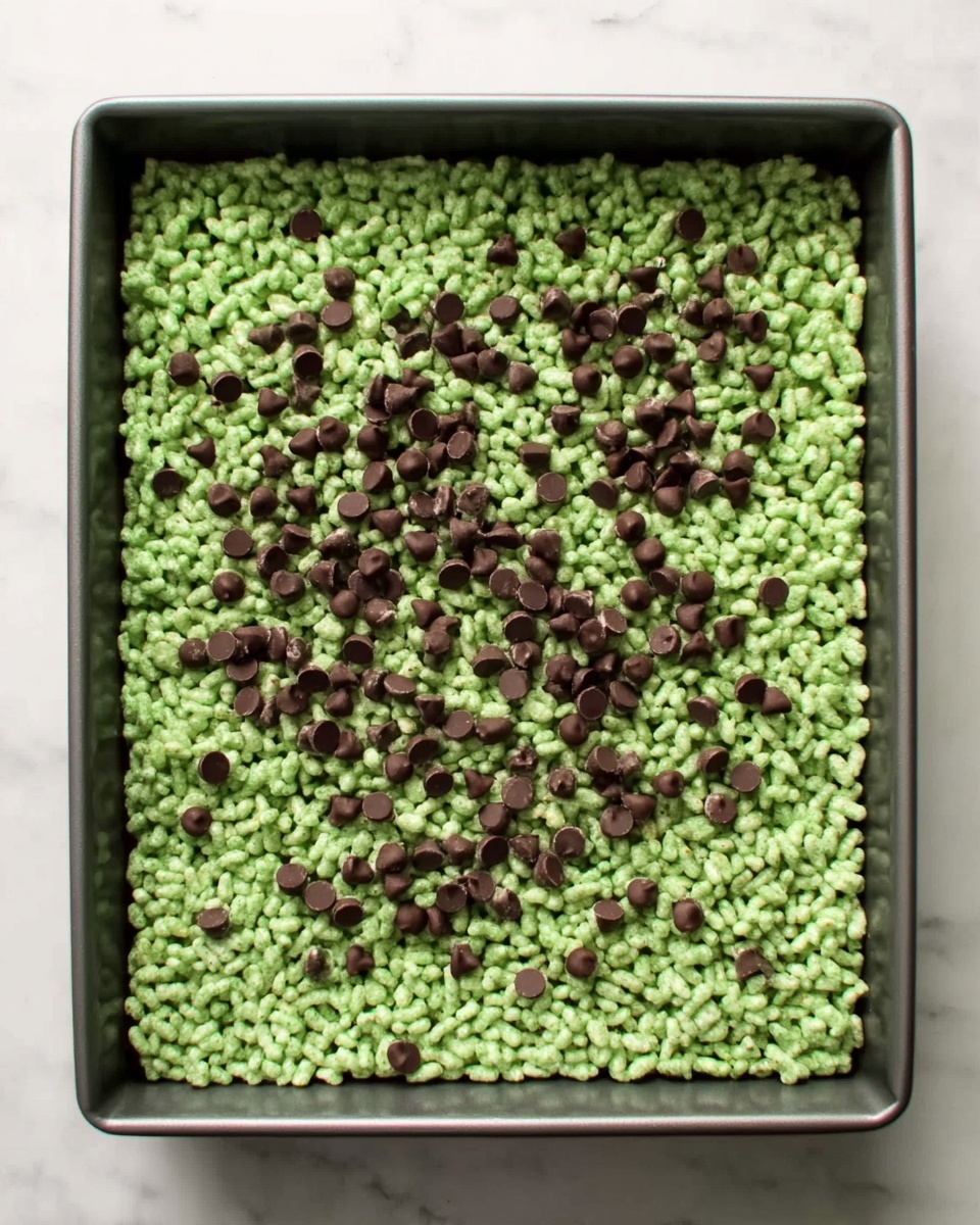 A square baking pan filled with a single thick layer of light green crispy rice treats mixed evenly with small dark chocolate chips scattered all over the top. The treat layer has a rough, bumpy texture from the rice cereal and chocolate chips. The pan is on a white marbled surface, showing the treat clearly from above. photo taken with an iphone --ar 4:5 --v 7