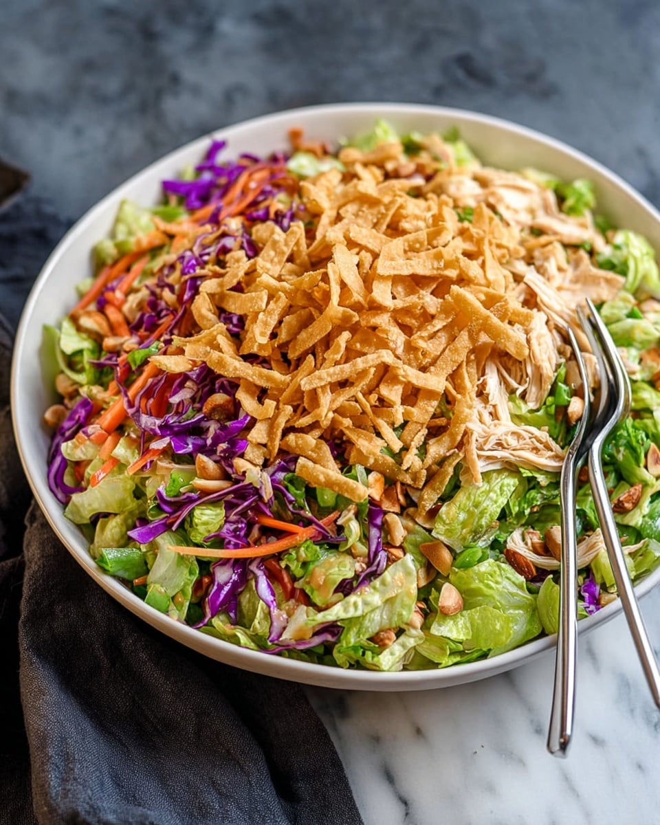 A large white bowl filled with a colorful layered salad is placed on a white marbled surface. The bottom layer consists of chopped green lettuce mixed with shredded orange carrots and purple cabbage. The middle layer shows sliced almonds and shredded light brown chicken spread evenly. The top layer has a heap of light golden fried wonton strips scattered above, giving a crunchy texture. The salad is vibrant with a mix of green, orange, purple, and light brown colors, with silver forks placed inside the bowl for serving. Photo taken with an iphone --ar 4:5 --v 7