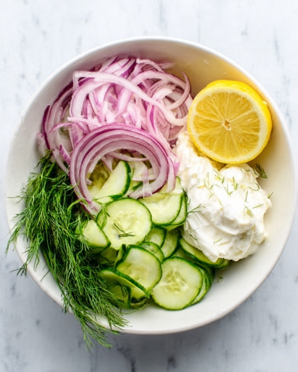 A white bowl on a white marbled surface holds a fresh and colorful salad with four main layers: thinly sliced red onions on the top left, bright yellow lemon wedge at the top right, a creamy white dollop of dressing or sauce to the right side, thin cucumber slices layered in the center and bottom right, and fresh green dill placed at the bottom left. The textures range from smooth and creamy in the dressing to crisp and fresh in the vegetables, creating a clean and bright look. photo taken with an iphone --ar 4:5 --v 7