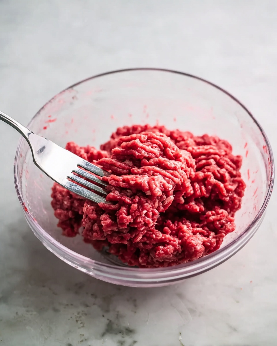 A clear plastic bowl sits on a white marbled surface, holding bright red raw ground meat clumped together in the center. A silver fork is stuck into the meat, angled slightly to the left, showing some strands of meat wrapped around the fork's tines. The texture of the meat is coarse, with visible small pieces grouped tightly, and the background is softly blurred. photo taken with an iphone --ar 4:5 --v 7