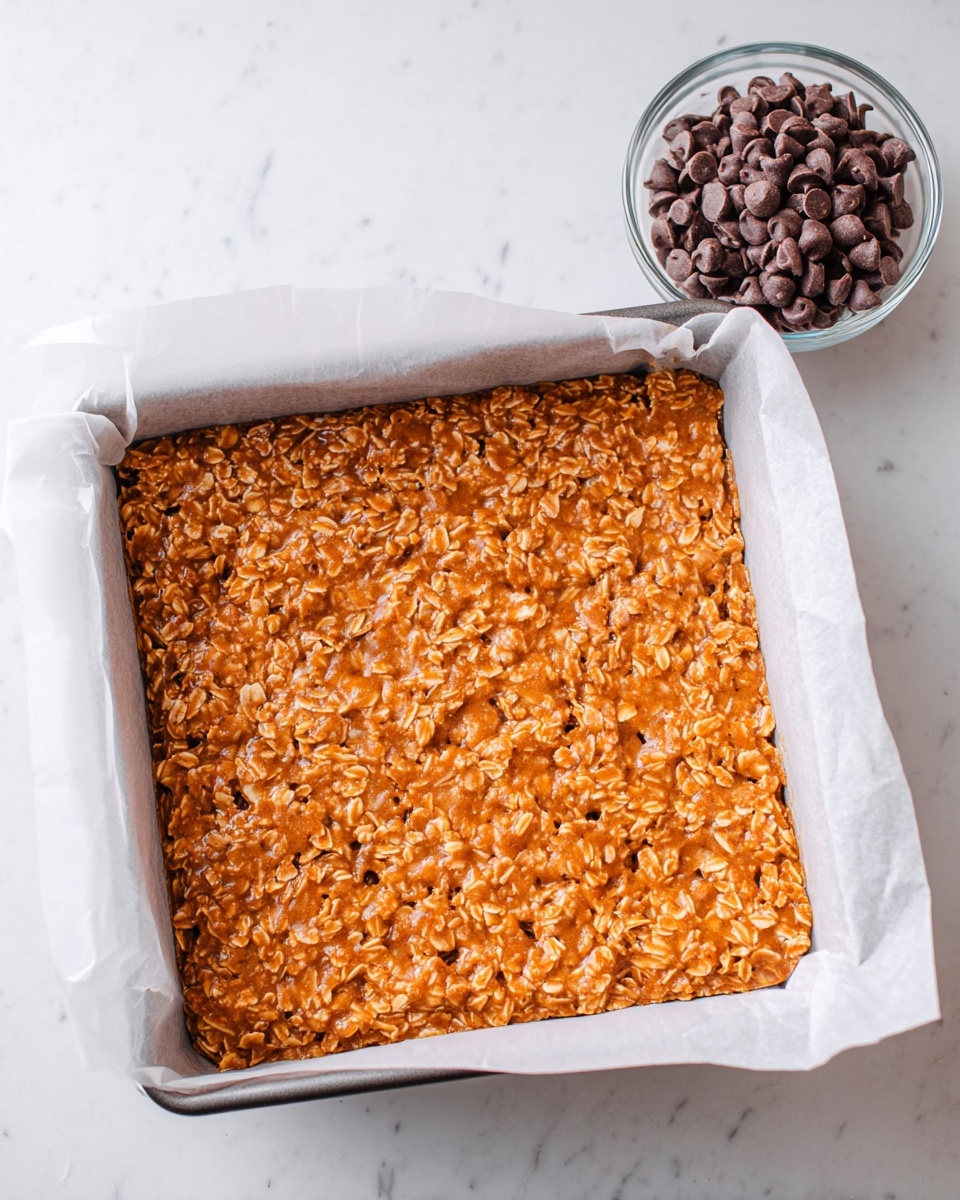 A square baking pan lined with white parchment paper holds a single thick layer of orange-brown mixture that looks sticky and textured with visible oats or rice flakes throughout. Next to the pan on a white marbled surface is a small clear glass bowl filled with dark brown chocolate chips. The lighting is bright and natural, showing the shine on the mixture and the smooth surface of the chocolate chips. photo taken with an iphone --ar 4:5 --v 7