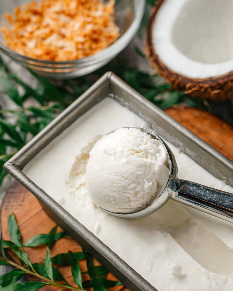 A scoop of smooth, creamy white ice cream is held in a shiny metal ice cream scoop, resting in a rectangular metal container filled with the same white ice cream. In the background, there is a half coconut shell showing its white inside, and a clear glass bowl filled with golden toasted coconut flakes. The setting includes green leaves and a wooden surface beneath the container, all creating a natural and fresh look. photo taken with an iphone --ar 4:5 --v 7