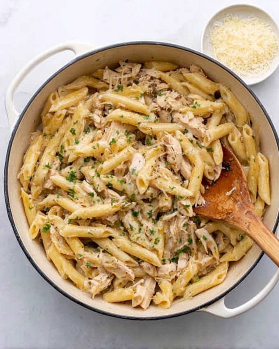 The image shows a white pot filled with cooked penne pasta mixed with shredded pieces of light brown chicken and small bits of green herbs scattered on top. The pasta looks creamy with a light sauce that coats all pieces evenly. A wooden spoon is partly inside the pot, lifting some pasta. On the side, there is a small white bowl with grated cheese. The pot is placed on a white marble surface. photo taken with an iphone --ar 4:5 --v 7