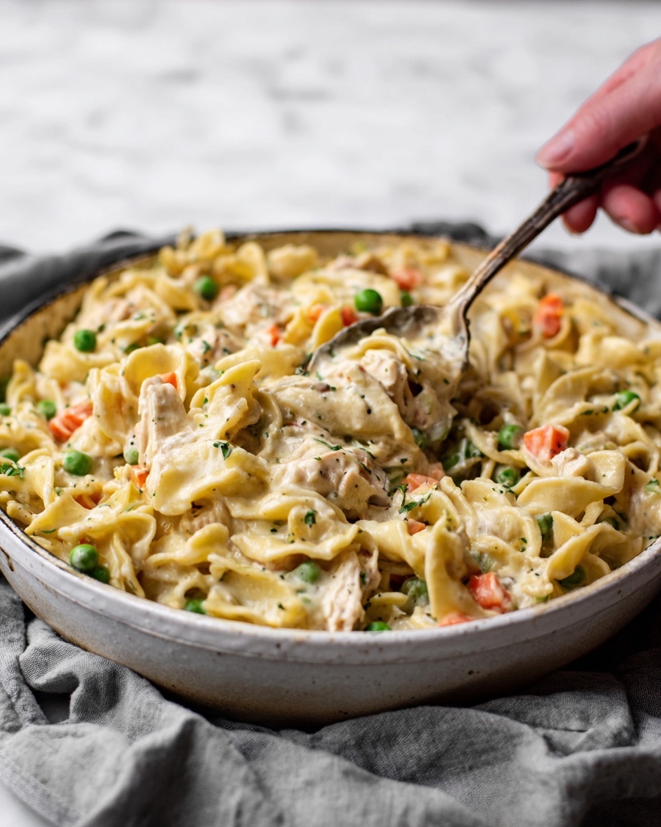 The image shows a close-up of a white bowl filled with creamy chicken noodle casserole. The dish has wide egg noodles mixed with small pieces of chicken and colorful bits of peas and carrots. The sauce looks thick and creamy, covering all the ingredients evenly. A woman's hand is using a spoon to scoop some of the casserole from the bowl. The background has a white marbled texture with a soft gray cloth next to the bowl. photo taken with an iphone --ar 4:5 --v 7