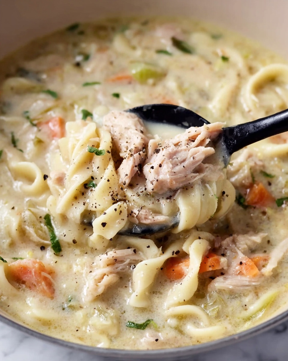 The image shows a creamy soup filled with thick, white noodles that twist through the dish, making up one main layer. Mixed in are pieces of soft, cooked meat with a light pink and beige color spread evenly throughout. Orange carrot chunks and small green herb leaves are scattered around within the soup's pale cream-colored broth that looks smooth and thick. There is a black spoon lifting part of the soup and meat, with tiny black pepper flakes sprinkled on top and some herbs adding a little bit of color contrast. The dish sits on a white marbled surface photo taken with an iphone --ar 4:5 --v 7