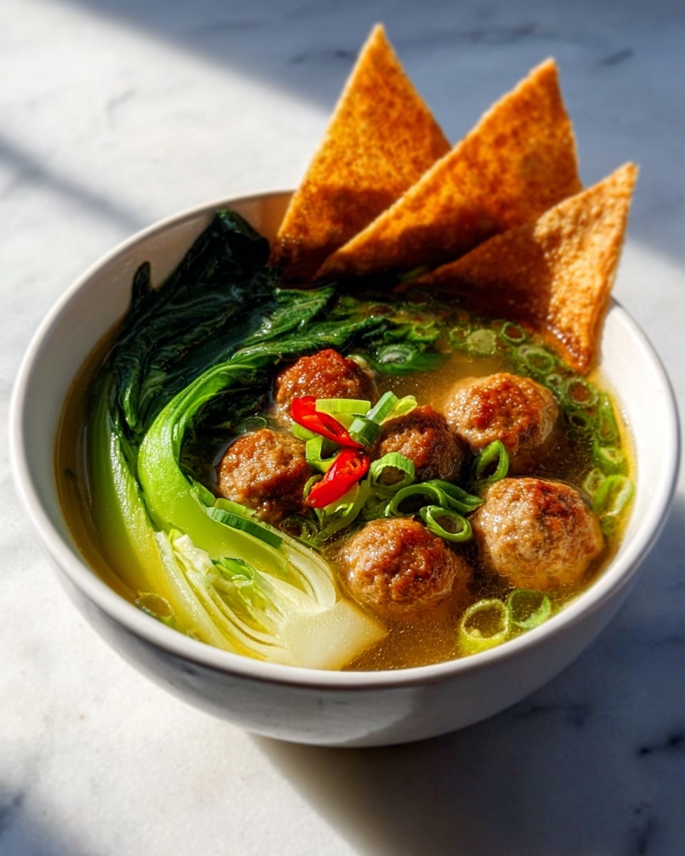 A white bowl filled with a clear broth containing several round, brown meatballs floating on top. Bright green bok choy is placed on one side of the bowl, with its leaves extending outward and its base submerged in the broth. Thin slices of green onion and small red chili pieces are scattered over the meatballs and broth, adding bursts of color. Three golden-brown crispy wonton chips are standing vertically on one edge of the bowl. The bowl is placed on a white marbled surface with natural light highlighting the textures. Photo taken with an iphone --ar 4:5 --v 7