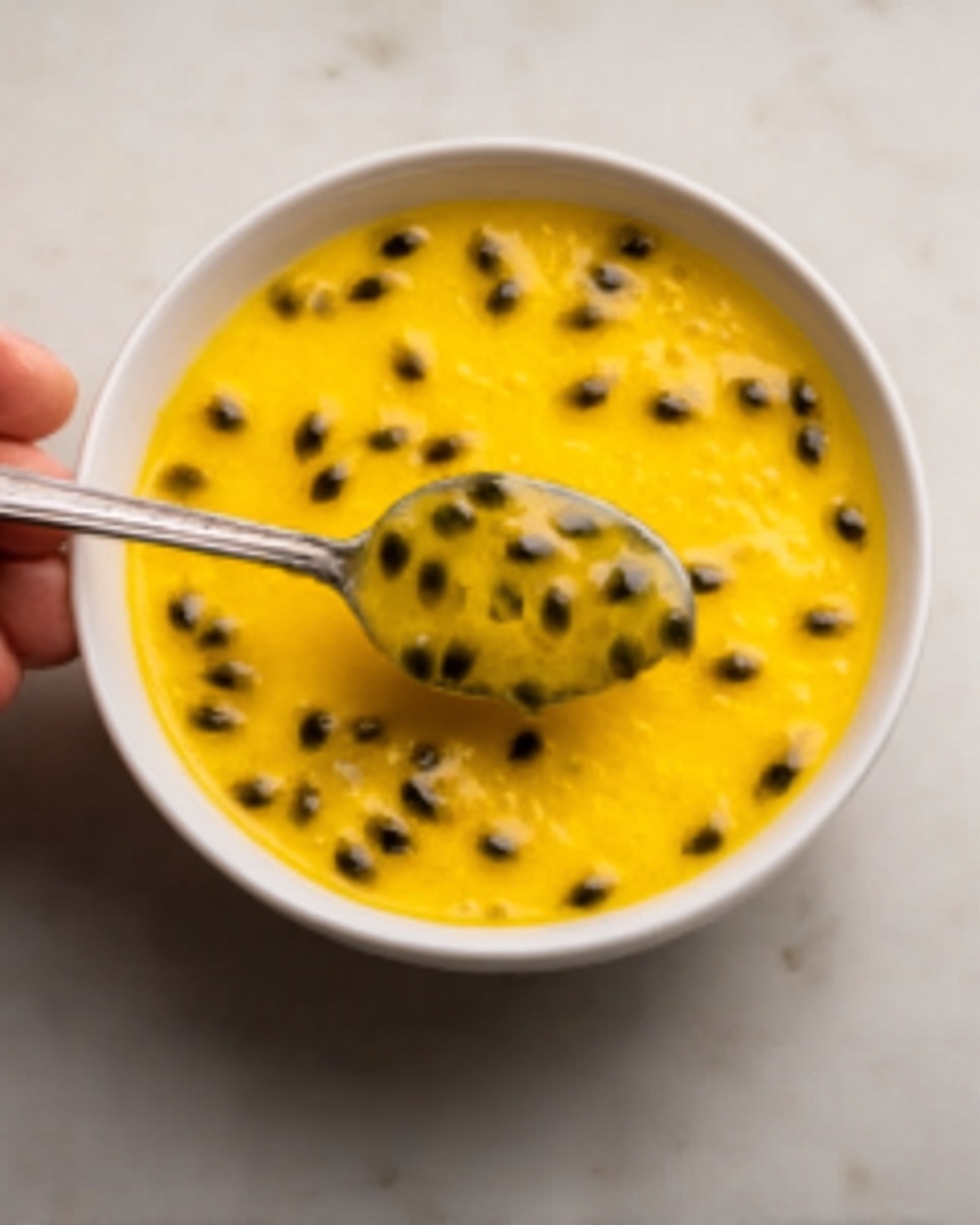 A white bowl filled with bright yellow passion fruit juice topped with black passion fruit seeds spread evenly across the surface. A woman's hand holding a silver spoon scoops up some of the juice with seeds, showing the smooth and slightly thick texture. The bowl sits on a white marbled surface, giving a clean and simple look. photo taken with an iphone --ar 4:5 --v 7