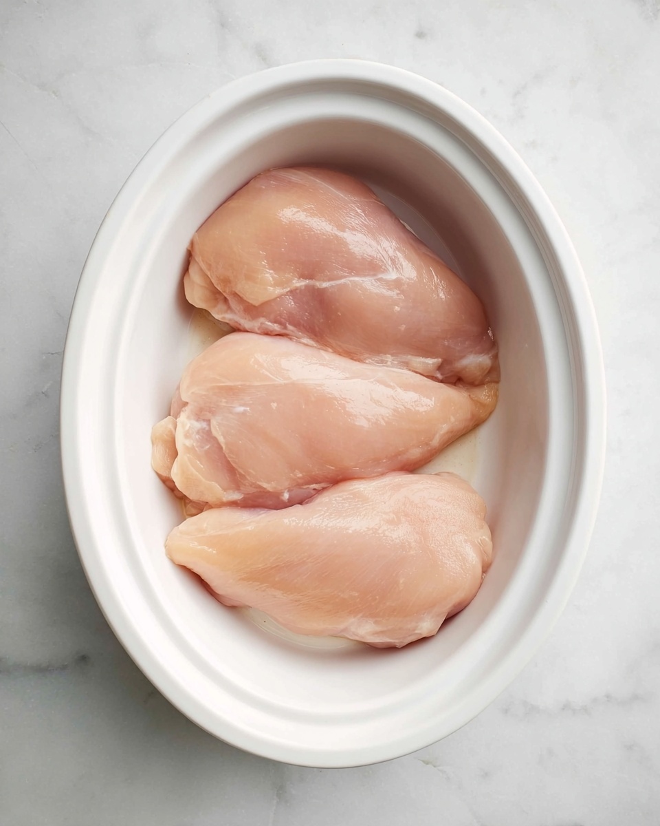 Three raw chicken fillets with smooth, pale pink skin lie close together in a deep white oval dish, which rests on a white marbled surface. The fillets have a soft, moist texture with slight sheen, filling the bottom of the dish evenly. The scene is lit softly from above, showing the natural details of the chicken skin and the clean, simple look of the dish and background. photo taken with an iphone --ar 4:5 --v 7