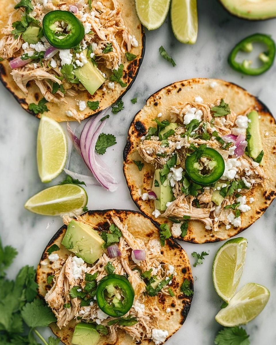 The image shows three small toasted tortillas on a white marbled surface, each topped with shredded light brown cooked chicken. On the chicken, there are small pieces of white cheese and tiny bits of chopped red onion and green cilantro. Each tortilla also has two or three slices of bright green jalapeño and a few chunks of pale green avocado. Lime wedges with a fresh pale green inside are placed around the tortillas along with some cilantro leaves and slices of red onion. The tortillas are slightly charred on the edges and the overall look is colorful and fresh. Photo taken with an iphone --ar 4:5 --v 7