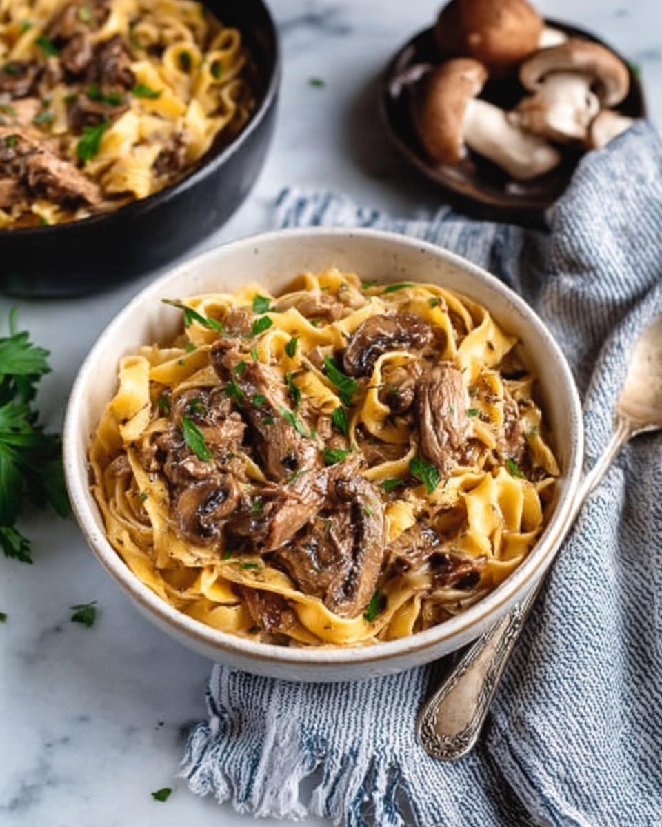 A white bowl filled with creamy fettuccine pasta layered with tender browned chicken pieces and sliced mushrooms. The sauce is light brown and coats the pasta evenly, with some green parsley leaves scattered on top for color. Next to the bowl on a white marbled surface is a spoon and a folded linen cloth with blue and white stripes. In the background, there's another bowl with more mushrooms. A woman's hand is reaching toward the bowl in the foreground. Photo taken with an iphone --ar 4:5 --v 7