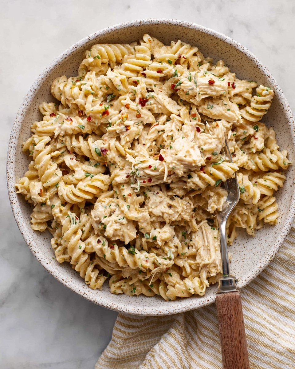 A white speckled bowl filled with creamy pasta and shredded chicken mixed together, the pasta spirals coated well in a light beige sauce with small green herb pieces and red pepper flakes sprinkled on top. The texture is rich and smooth with tender shredded chicken blended evenly throughout. A fork with a wooden handle rests inside the bowl, partially lifting some pasta and chicken. The bowl is set on a white marbled surface with a beige and white striped cloth partially visible underneath. photo taken with an iphone --ar 4:5 --v 7