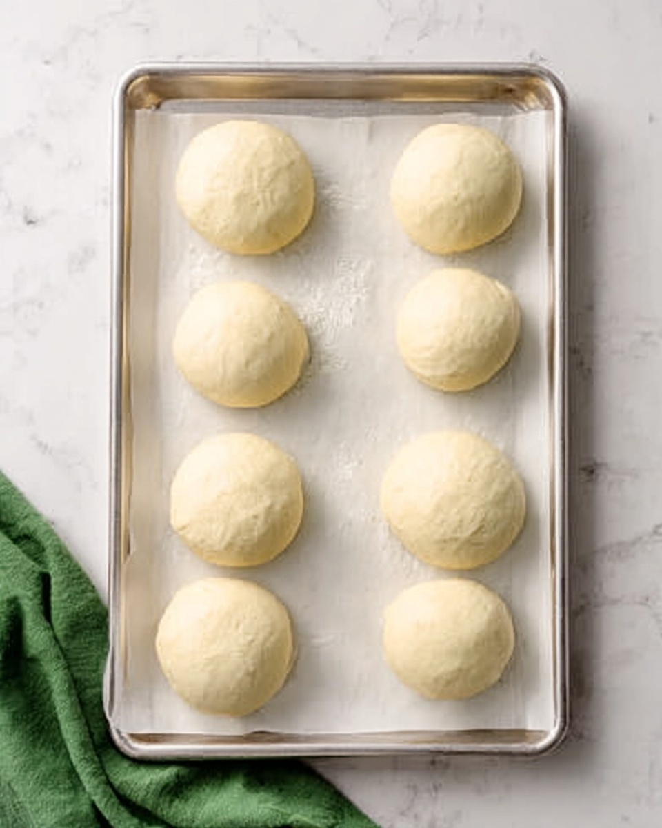The image shows eight dough balls placed evenly on a silver baking tray lined with white parchment paper. The dough balls are smooth and round with a pale cream color and soft texture. The tray is set on a white marbled surface with a green cloth partially visible at the bottom left corner. The lighting is bright and natural, highlighting the gentle shadows around the dough balls. photo taken with an iphone --ar 4:5 --v 7