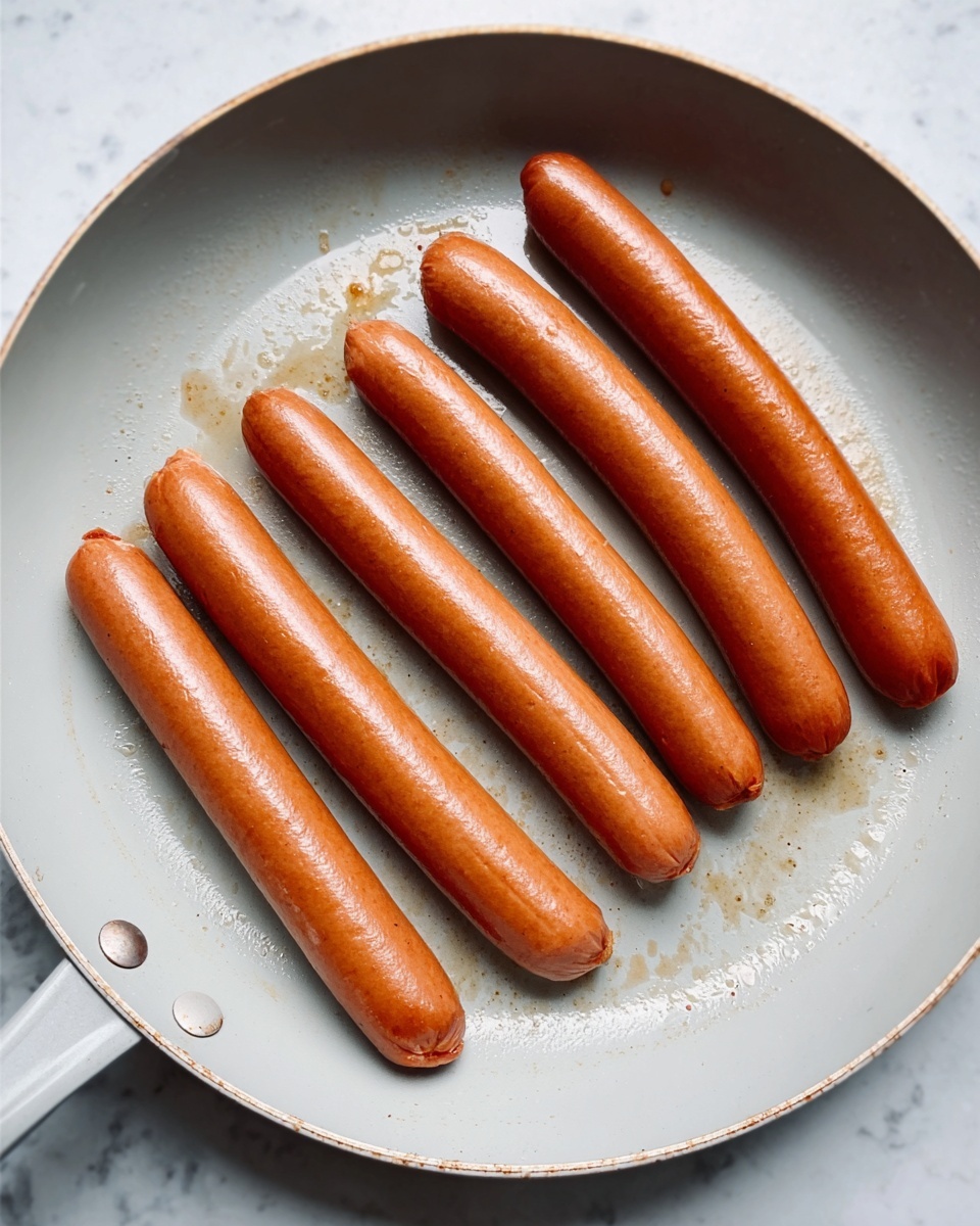 The image shows a white frying pan with six long, brown sausages lined up, evenly spaced. The pan has a smooth light gray surface with a little bit of oil showing where some sausages were moved, creating shiny spots. The sausages have a shiny, smooth texture with a slight browning on the tops. The background surface is a white marbled texture. Photo taken with an iphone --ar 4:5 --v 7