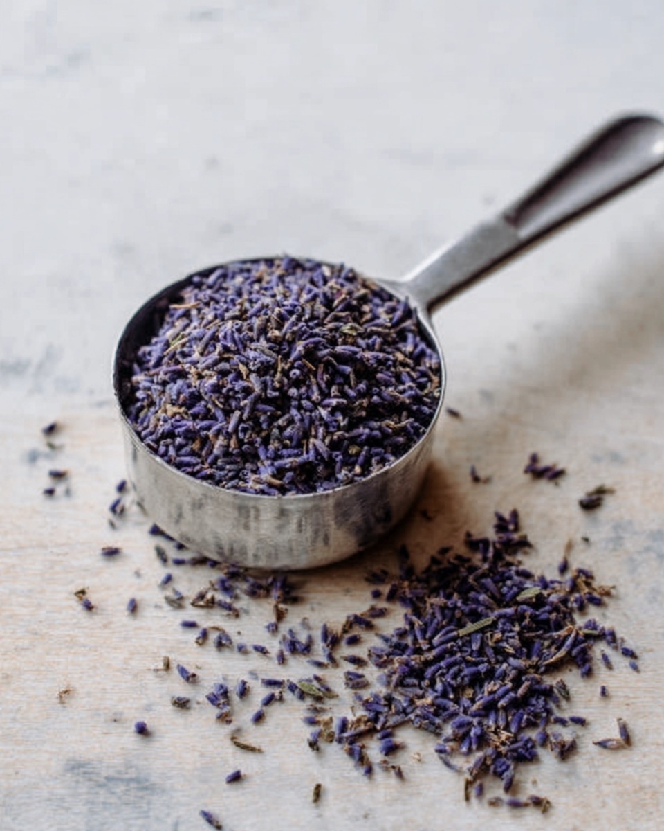 The image shows a small metal measuring cup filled to the top with dried lavender flowers. The lavender is dark purple with some small green stems mixed in, and the flowers have a fine, delicate texture. Some dried lavender petals have spilled out of the cup and scatter loosely on the light-colored wooden surface below. The metal cup has a smooth, shiny finish with a rounded handle extending to the right. The background surface is a white marbled texture. photo taken with an iphone --ar 4:5 --v 7