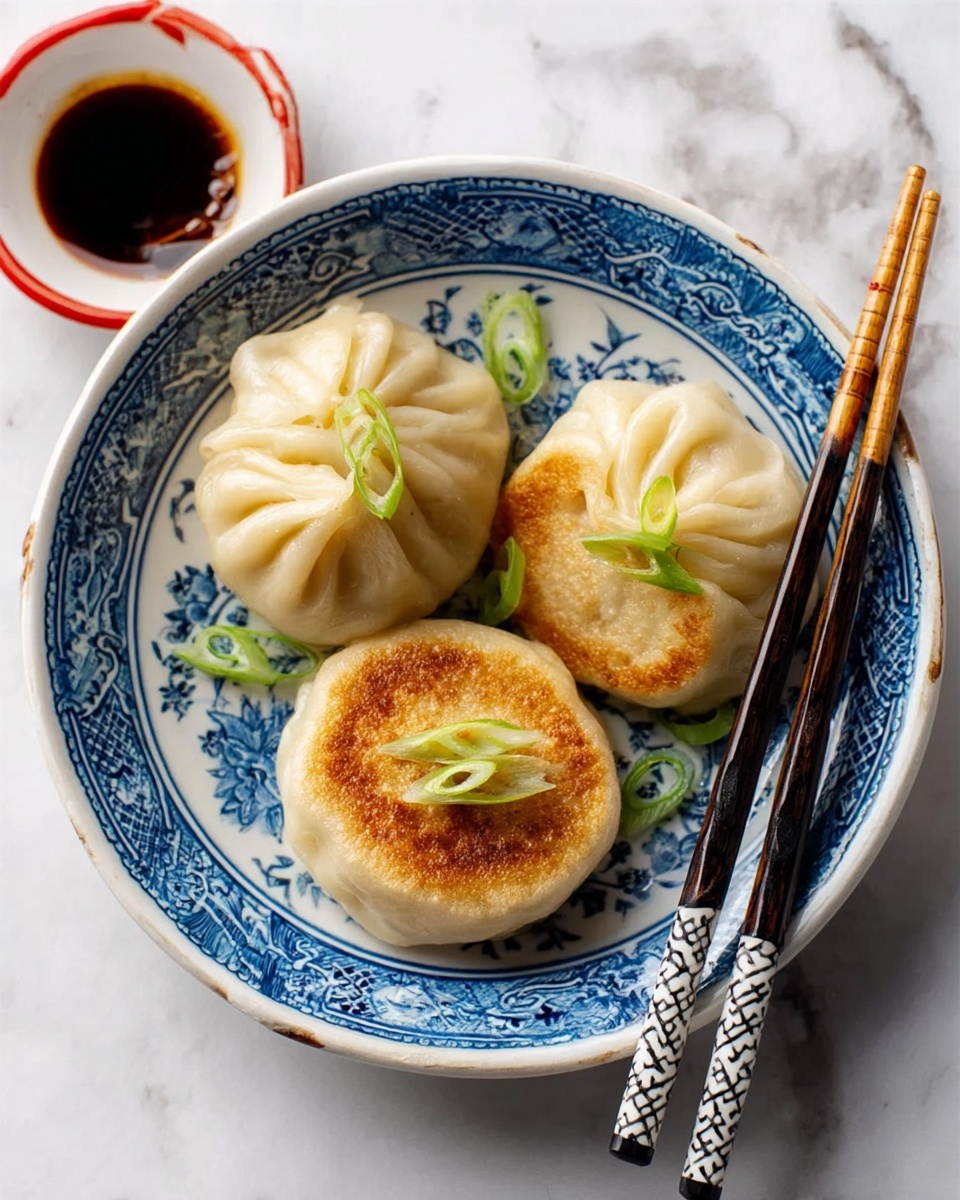 A white bowl with detailed blue patterns holds three round pan-fried dumplings with golden brown bottoms and pleated light beige tops. Each dumpling is topped with small green onion slices, and the bowl rests on a white marbled surface with a pair of black and white chopsticks nearby. A white bowl containing dark soy sauce with a red rim is partially visible next to the dumplings. photo taken with an iphone --ar 4:5 --v 7
