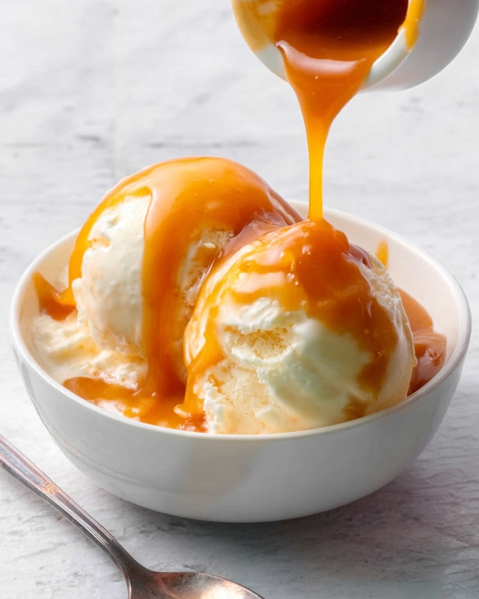 The image shows a white bowl with two round scoops of creamy vanilla ice cream inside. A rich, thick caramel sauce is being poured over the scoops from a small container held just above the bowl, covering the ice cream with a shiny, smooth drizzle. The bowl sits on a white marbled surface, and a silver spoon is placed next to it. The light is soft, bright, and natural, highlighting the creamy texture of the ice cream and the glossy caramel sauce. photo taken with an iphone --ar 4:5 --v 7