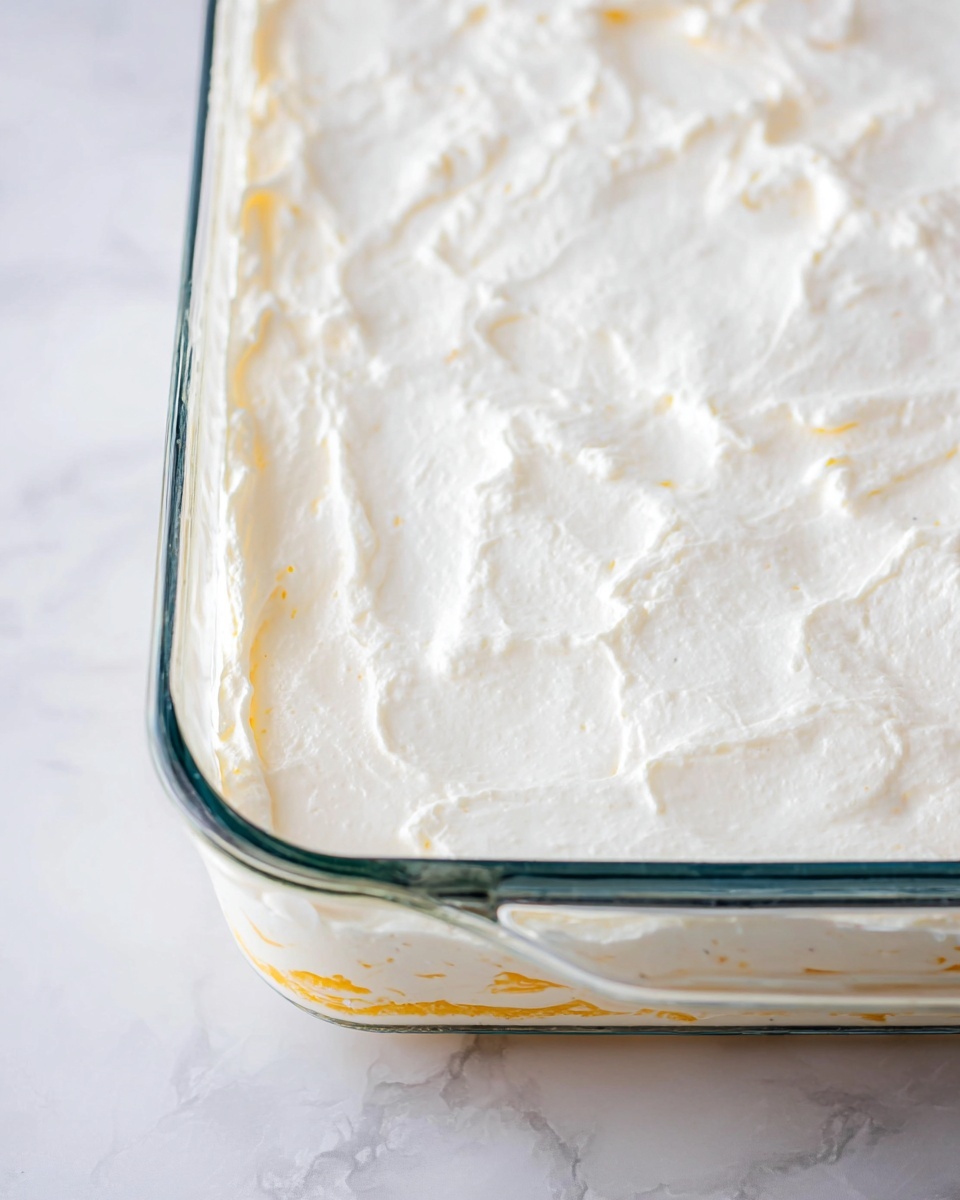The image shows a clear glass baking dish filled with a layered dessert or dish. The top layer is a thick, smooth white cream that has slight uneven textures and some small hints of yellow beneath it, showing that there are other layers or ingredients underneath. The dish is placed on a white marbled surface, which adds subtle texture to the background. The focus is on the creamy top layer and the edges of the clear dish, showing the thickness and smoothness of the cream layer. photo taken with an iphone --ar 4:5 --v 7