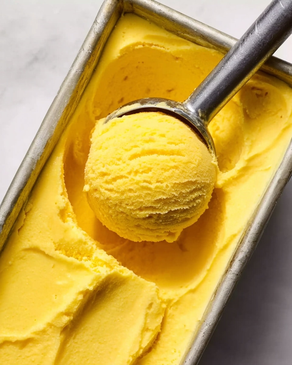 The image shows a close-up view of a metal ice cream scoop resting inside a rectangular metal container filled with smooth, bright yellow ice cream. The ice cream has a creamy texture and a shiny surface, with one round scoop lifted by the scoop, showing a dense and soft consistency. The container has slightly raised edges, and the scene is set against a white marbled background, enhancing the vivid yellow color of the ice cream. Photo taken with an iphone --ar 4:5 --v 7