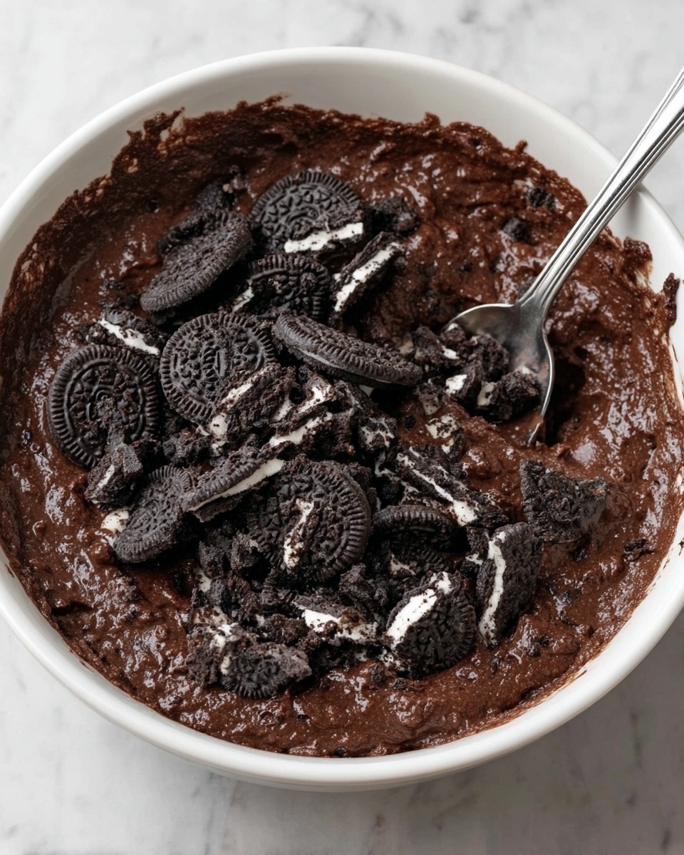 A white bowl filled with thick, dark brown chocolate batter that has a rich, slightly textured surface. On top, there is a generous layer of roughly chopped black and white sandwich cookies scattered unevenly, showing both the dark cookie parts and the white cream centers. A shiny silver spoon is partially immersed in the batter toward the right side of the bowl. The bowl is placed on a white marbled surface. photo taken with an iphone --ar 4:5 --v 7