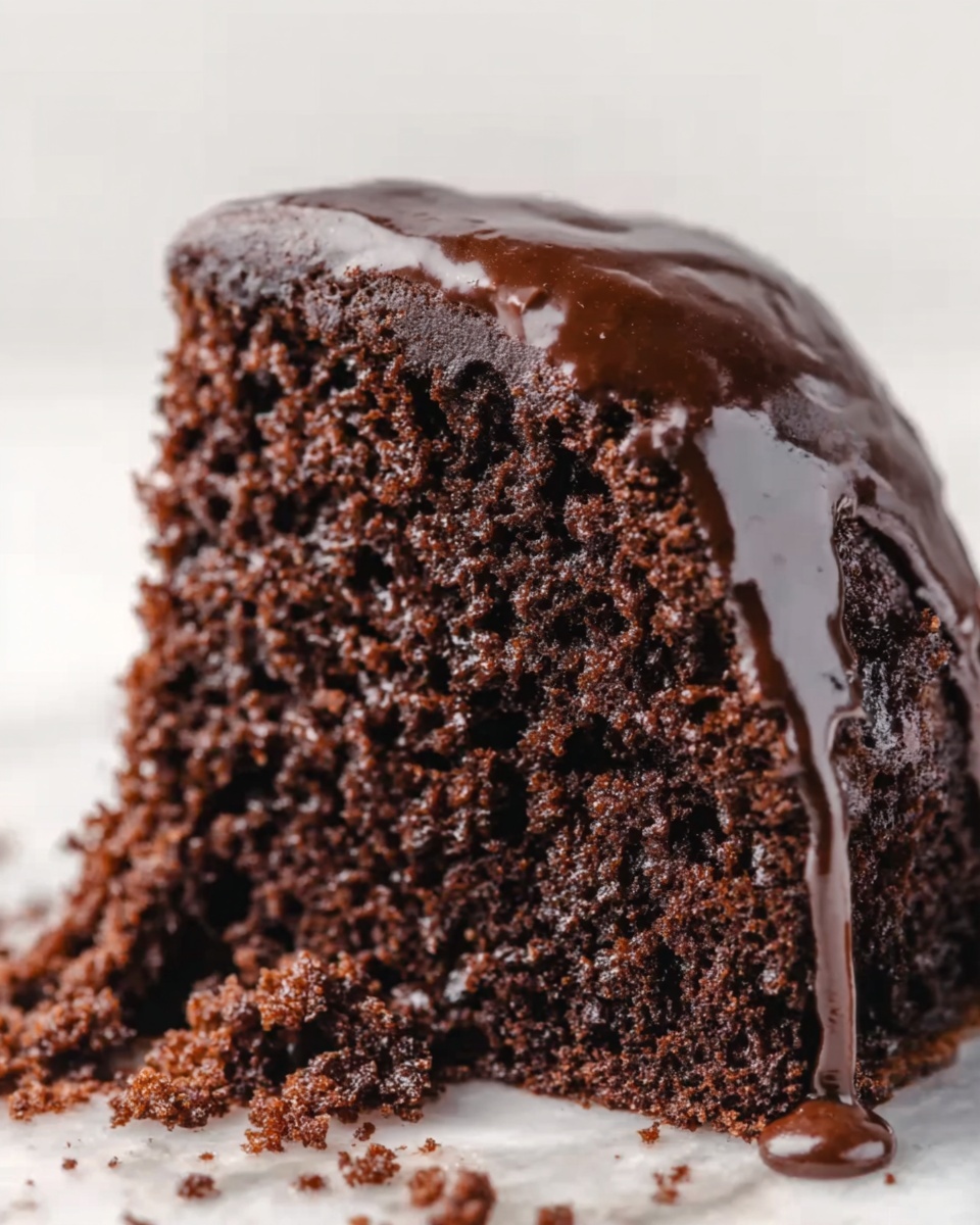 A close-up image of a rich, moist chocolate cake slice with one main thick layer. The top of the cake is covered with a smooth, shiny dark chocolate glaze that slightly drips on the sides. The crumb of the cake is dense and textured, showing small air holes throughout. The cake sits on a white marbled surface, and crumbs are scattered around the base. Photo taken with an iphone --ar 4:5 --v 7