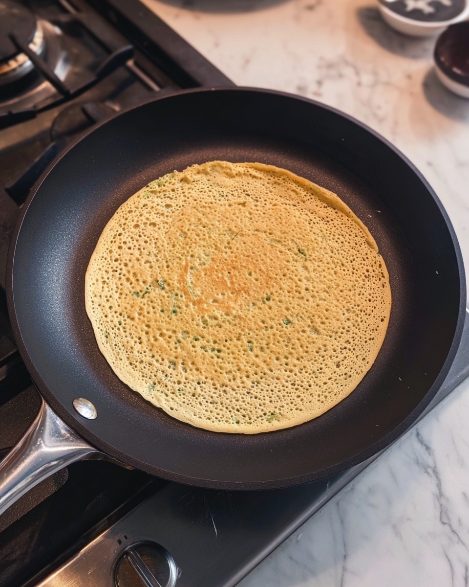 A single round crepe-like pancake with a light golden yellow color and small green flecks, sitting flat in the middle of a black frying pan on a gas stove. The pancake has a porous, slightly spongy texture with tiny holes covering its surface evenly. The pan has a smooth non-stick finish with a shiny silver handle visible at the bottom, and the stove's metal burners and control knob can be seen nearby. The background features a white marbled countertop with some kitchen items blurred in the top right corner. photo taken with an iphone --ar 4:5 --v 7