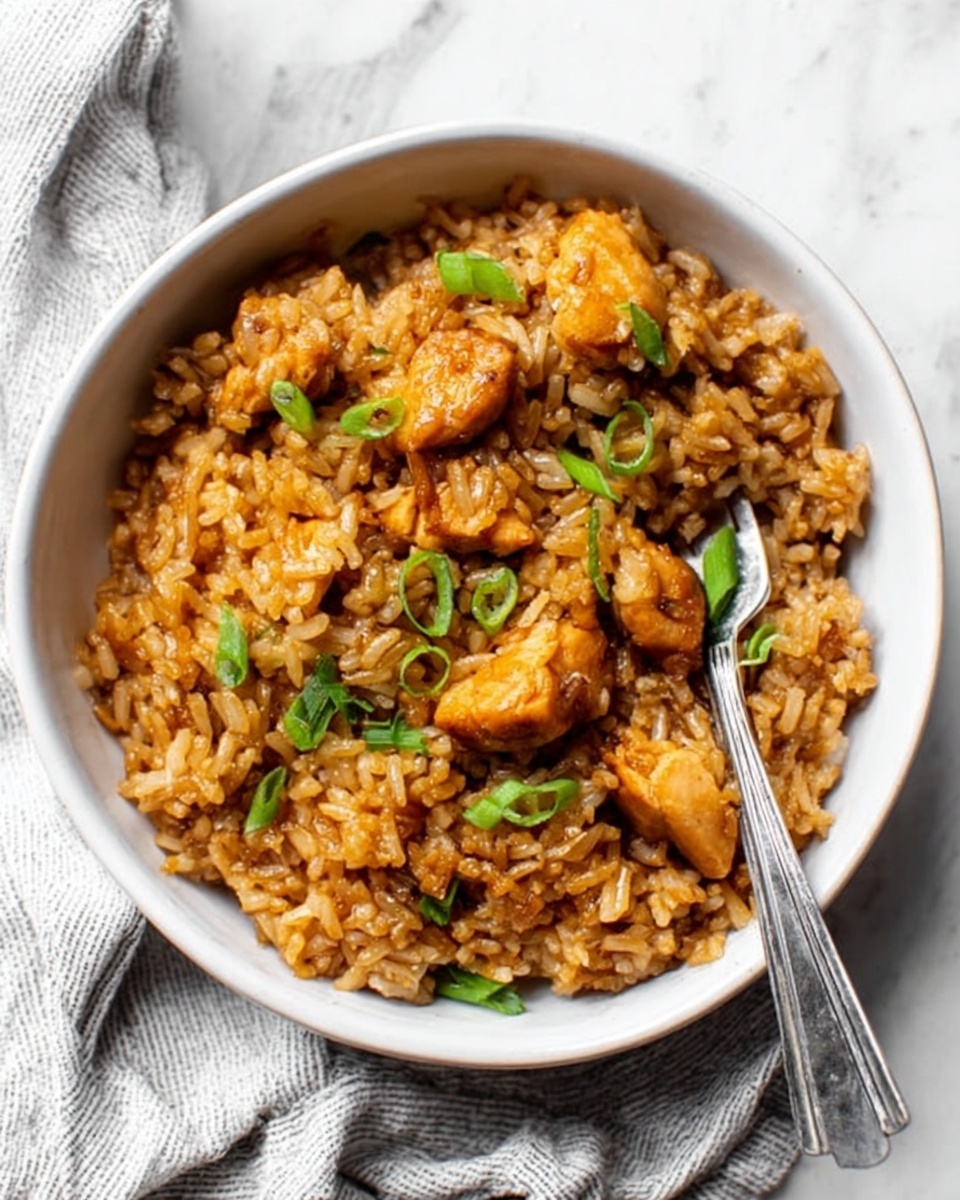 A white bowl filled with a thick mix of cooked rice and small pieces of golden brown chicken scattered evenly throughout. The rice looks soft and slightly sticky, with a rich, warm brown color from the sauce. Green sliced scallions are lightly sprinkled on top for a fresh touch. A silver fork rests inside the bowl on the right side, partially buried in the rice. The bowl sits on a white marbled surface with a soft, casual cloth partially visible underneath. Photo taken with an iphone --ar 4:5 --v 7