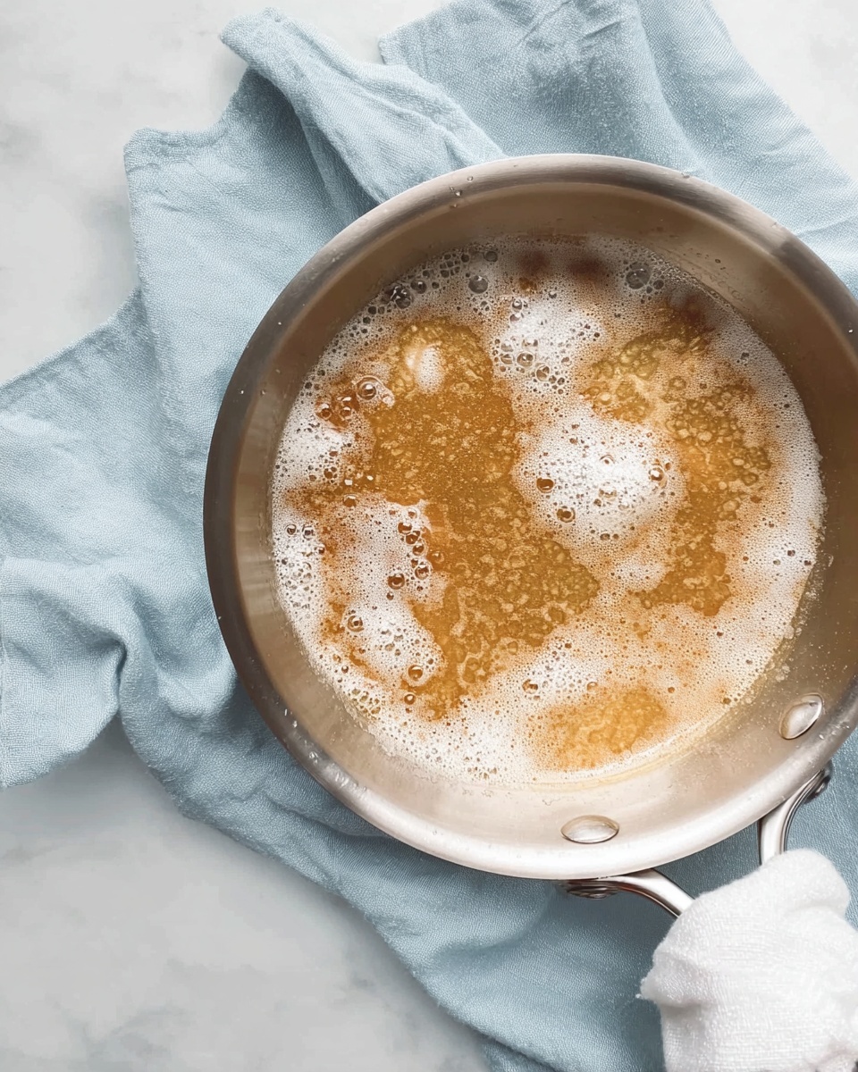 A large silver pan filled with a golden-brown liquid that has white foam patches scattered across the surface, showing a slightly bubbly texture. The pan rests on a soft light blue cloth, with a white cloth wrapped around the handle. The background is a white marbled surface, giving a clean and bright setting. Photo taken with an iphone --ar 4:5 --v 7